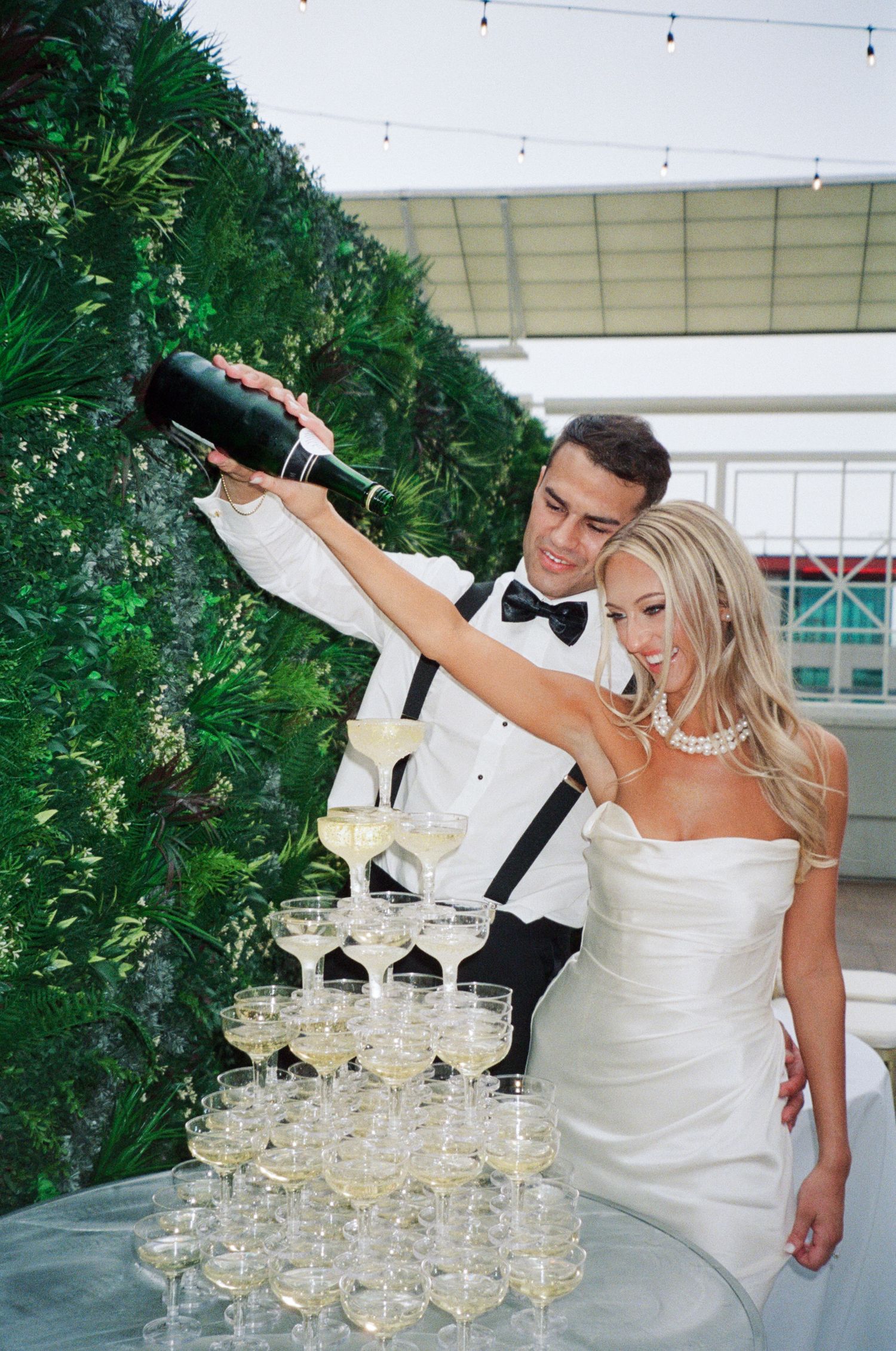 Bride and groom pouring champagne tower at wedding reception with green wall backdrop.