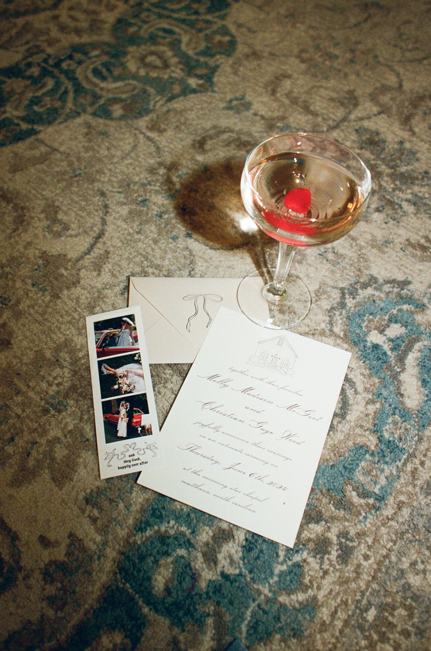 Wedding invitation, monogrammed envelope, photo booth strip and a pink cocktail in a coupe glass resting on an ornate blue patterned rug.