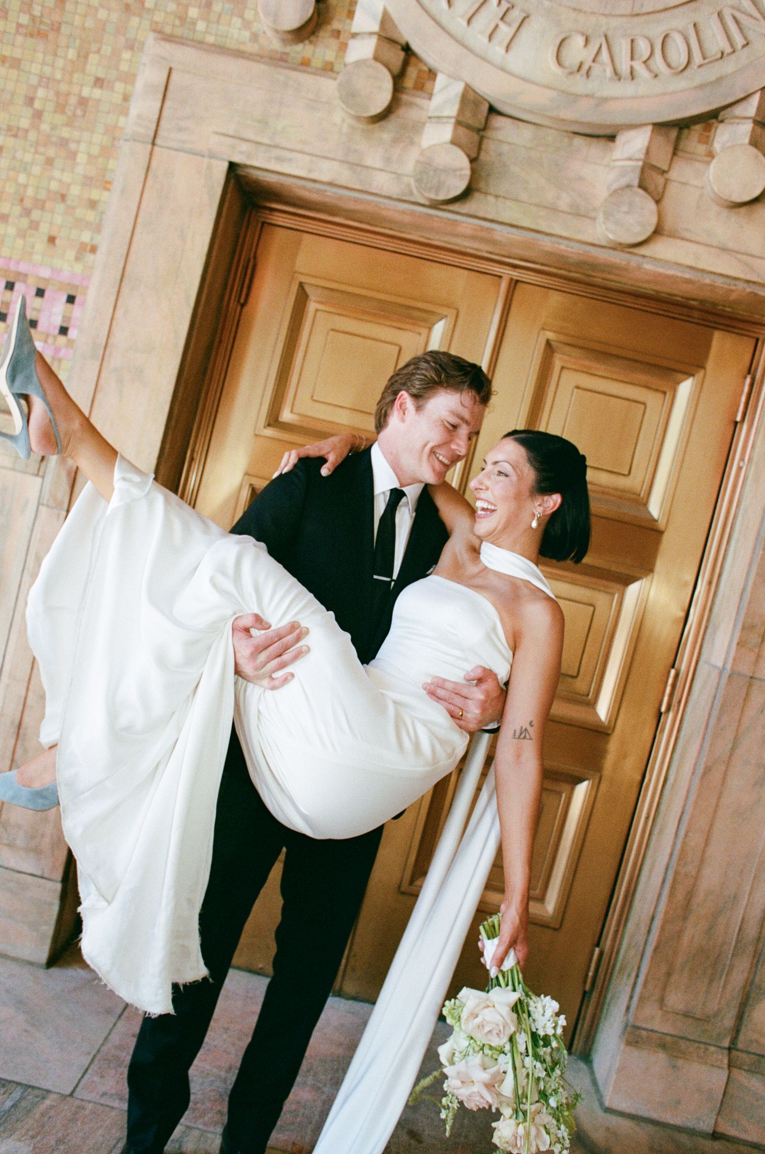 Smiling groom in a dark suit carries his laughing bride in a white wedding gown and bouquet in front of ornate golden doors.