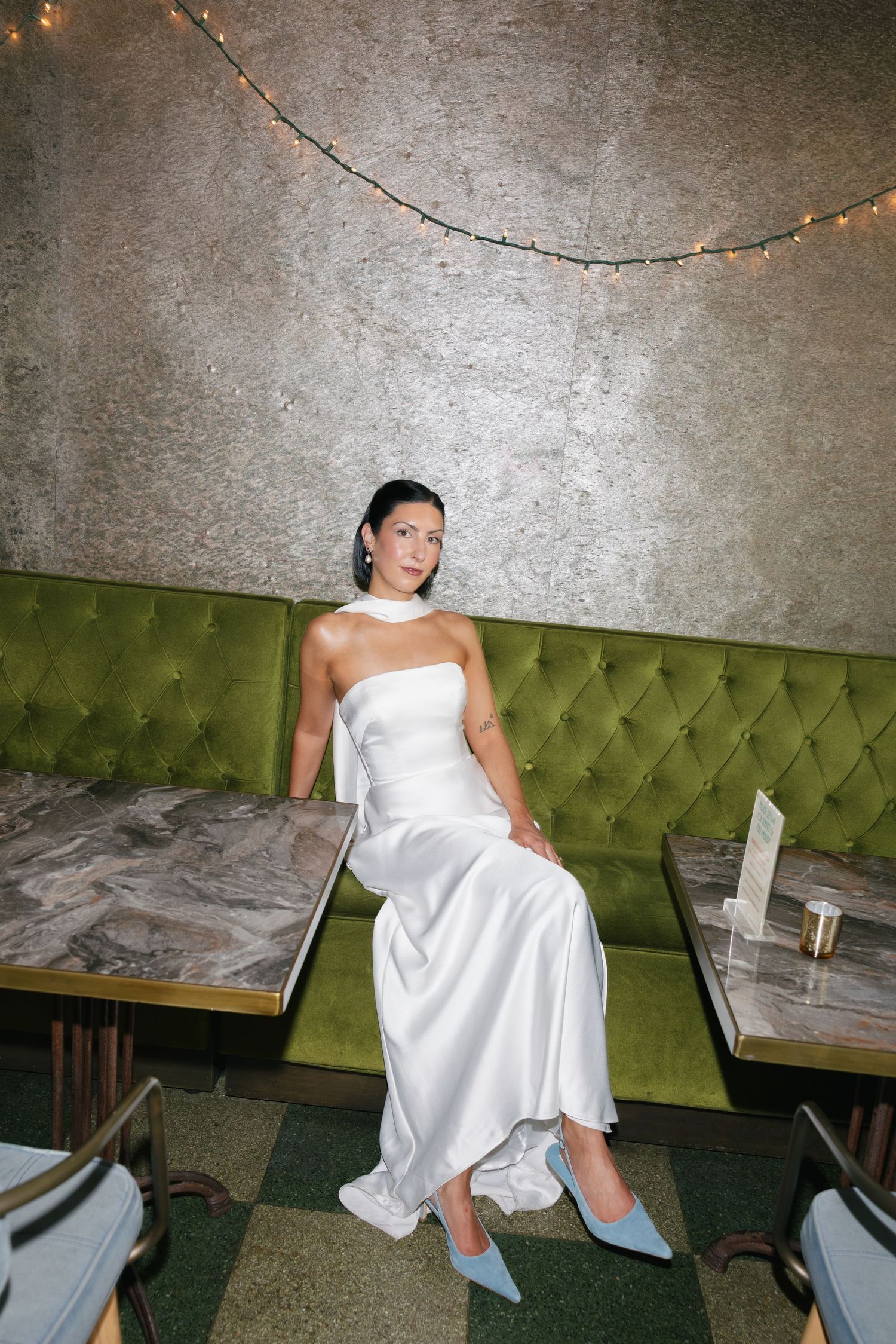 Woman in a white satin strapless gown and light blue pointed heels sits on a green tufted banquette in a restaurant with string lights.