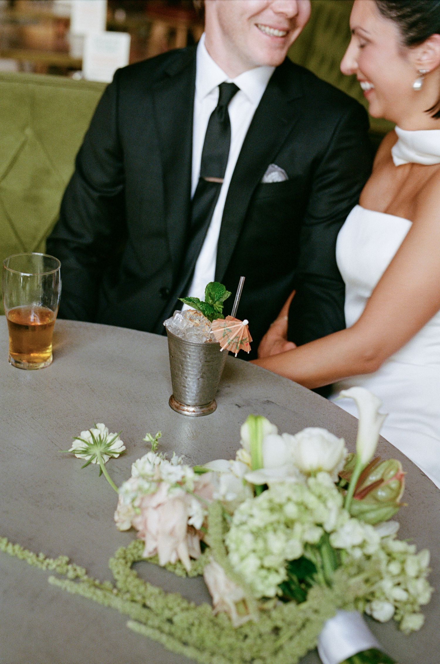 Smiling bride and groom sit at a round table with a white floral bouquet and a mint julep cocktail in a silver cup.