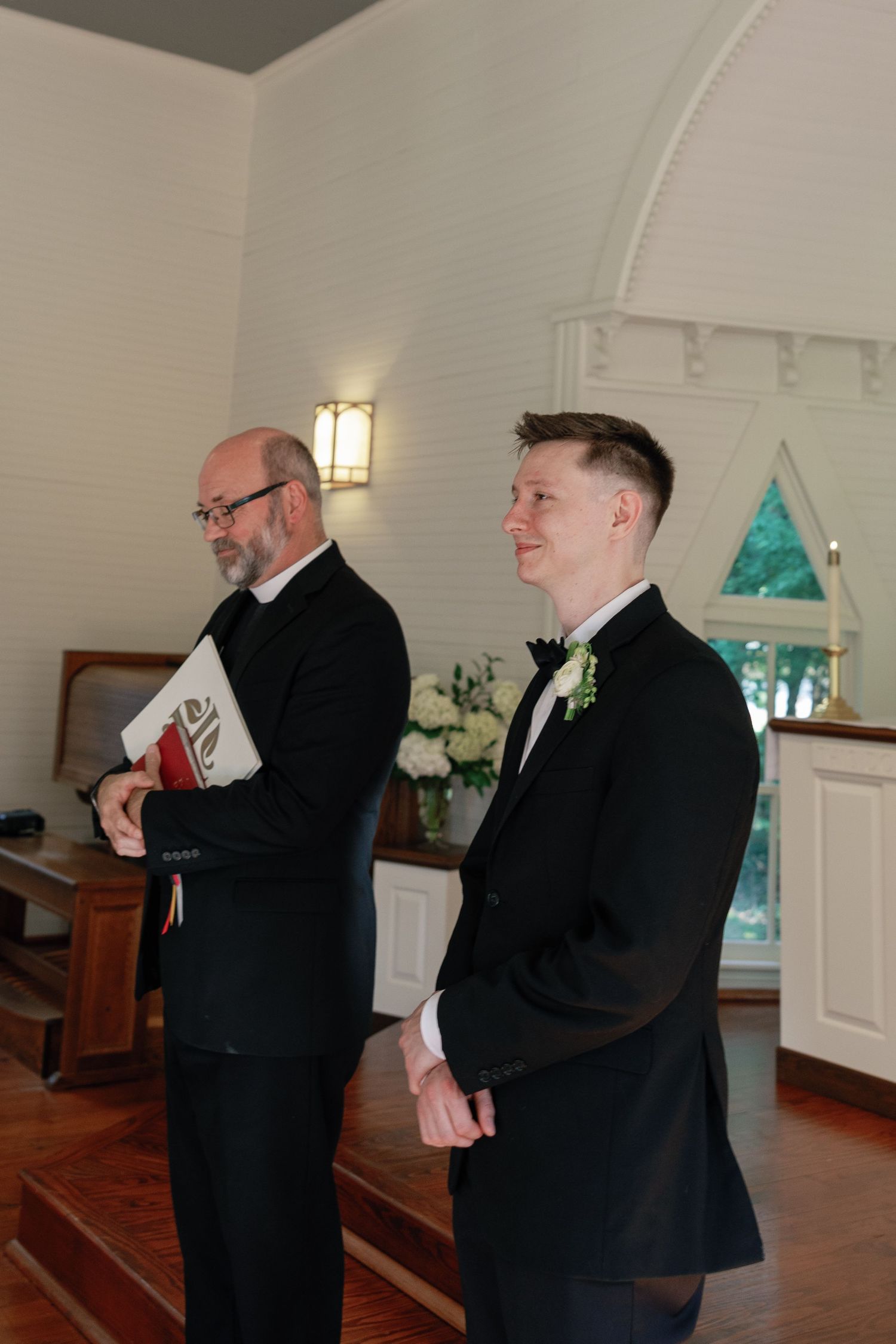 Smiling groom in a black suit with a floral boutonniere stands at the altar beside an officiant holding a book in a white wedding chapel.