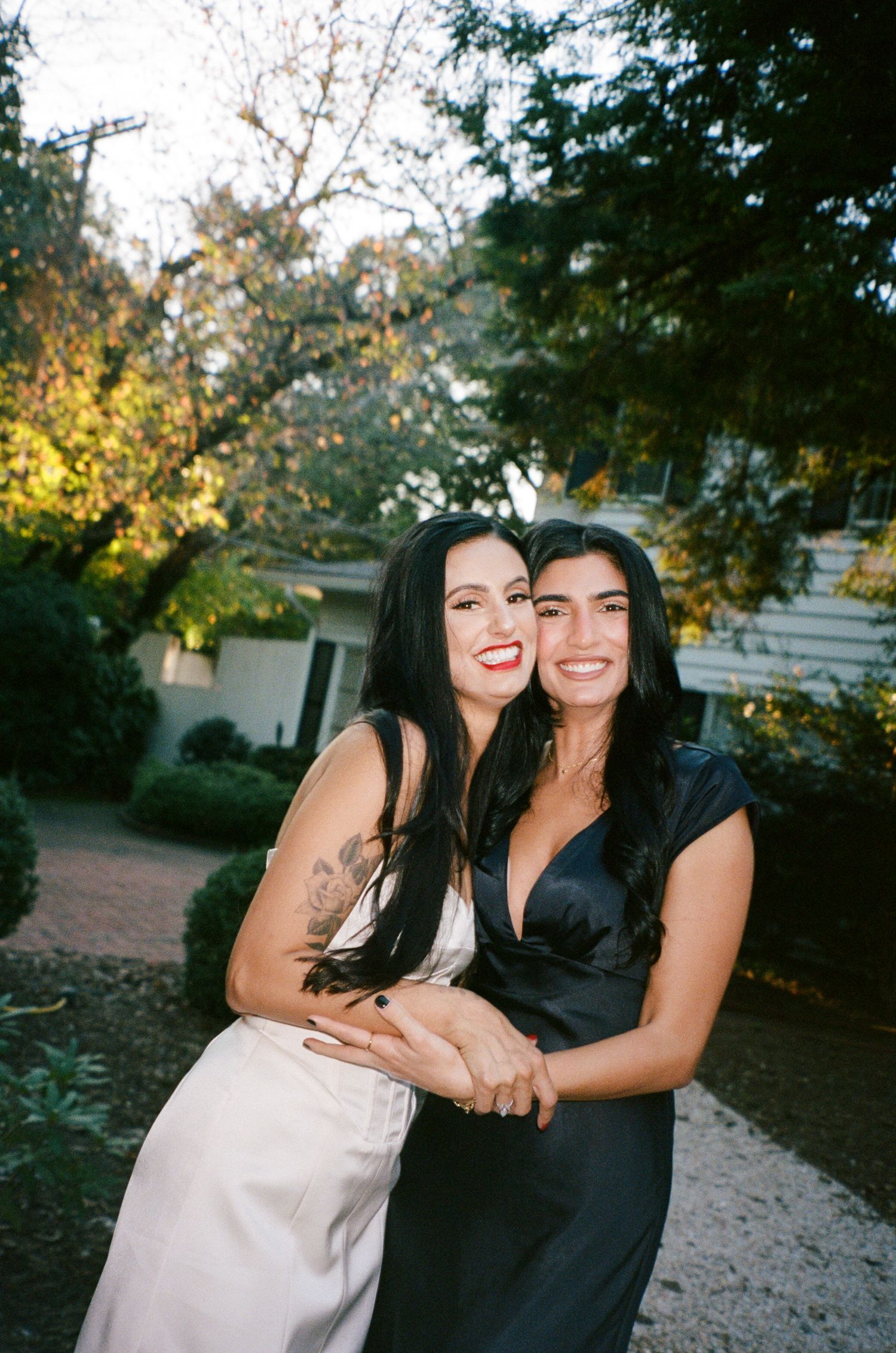 Two smiling women with long dark hair embrace outdoors, one in a white satin dress and one in a black dress, in front of trees and a house.