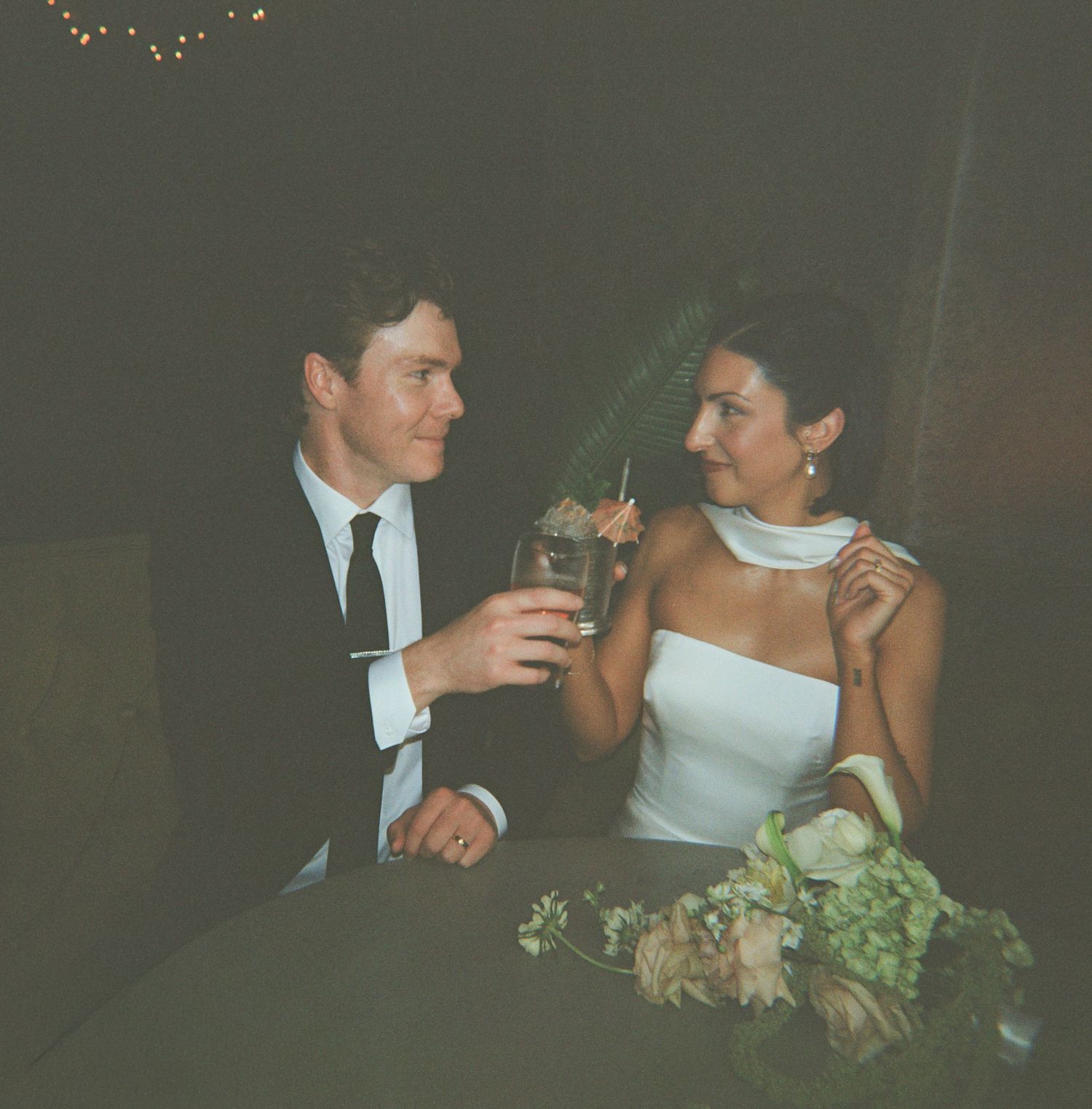 A smiling man in a dark suit and a woman in a white dress toast their drinks at a table with a flower bouquet at an intimate celebration.