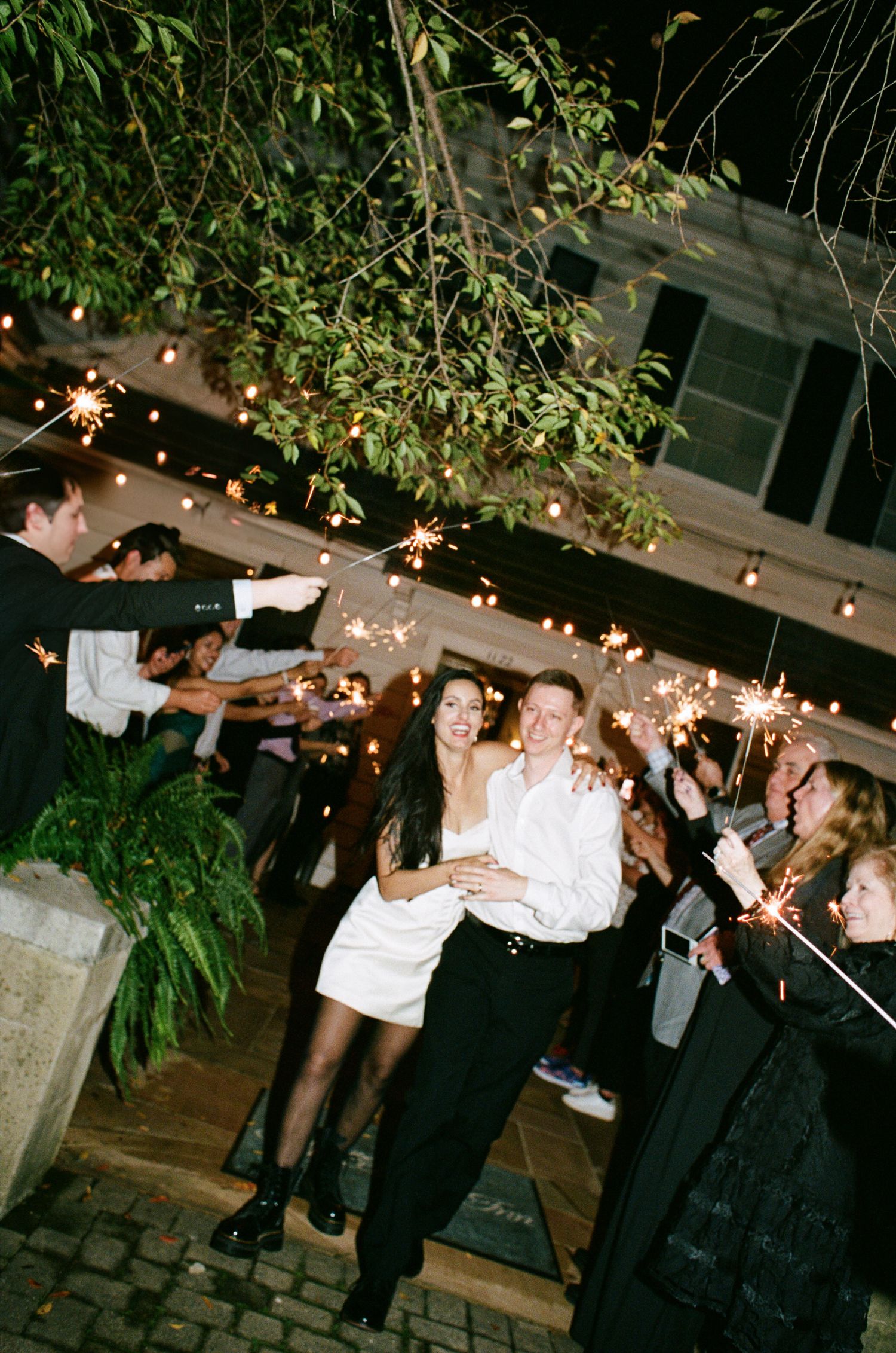 Bride and groom walk through guests holding sparklers during a nighttime wedding exit under string lights outside a historic building.