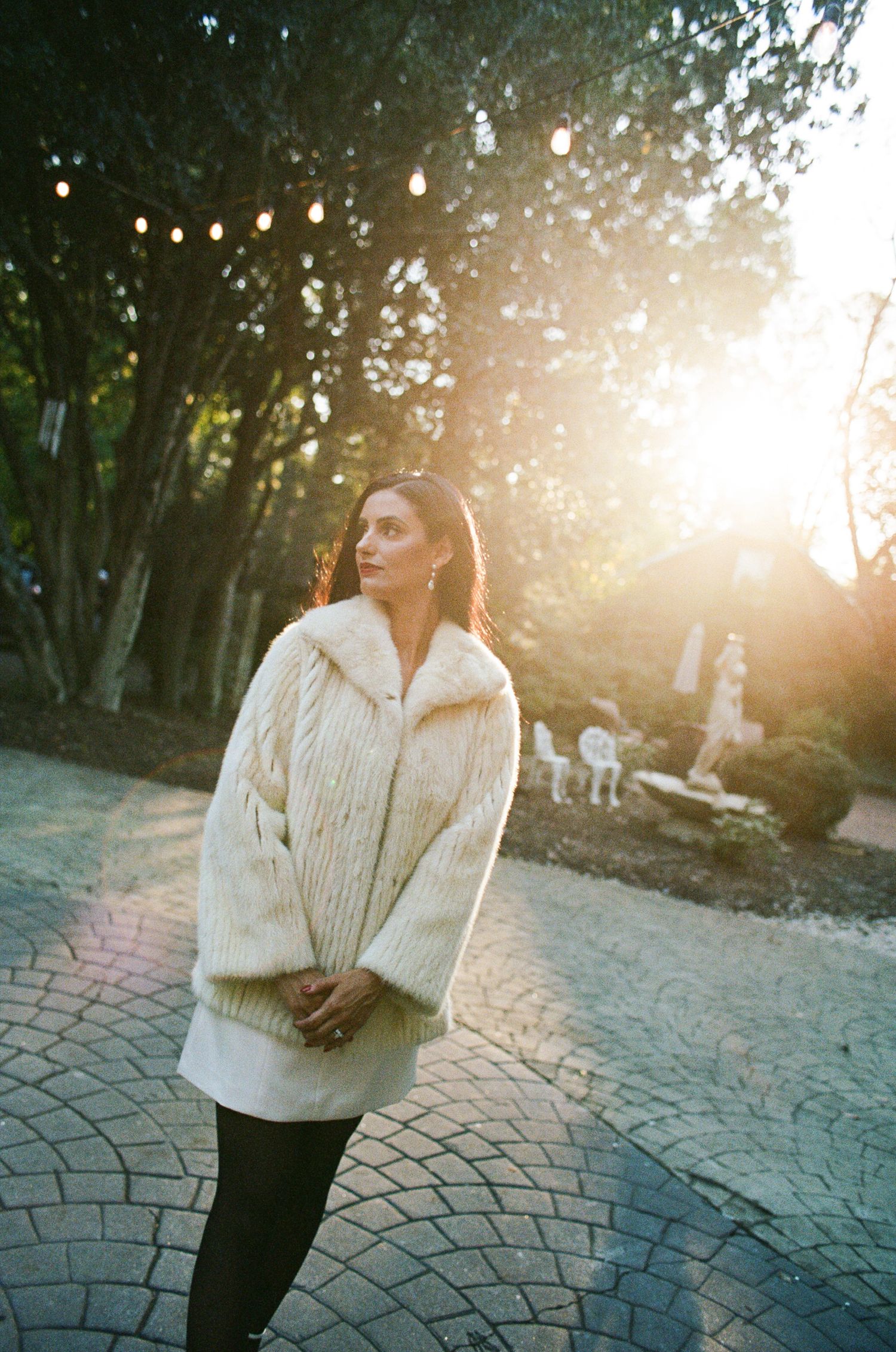 Woman in a cream cable-knit fur-collar coat stands on a cobblestone path at golden hour, with string lights and sun glowing behind her.