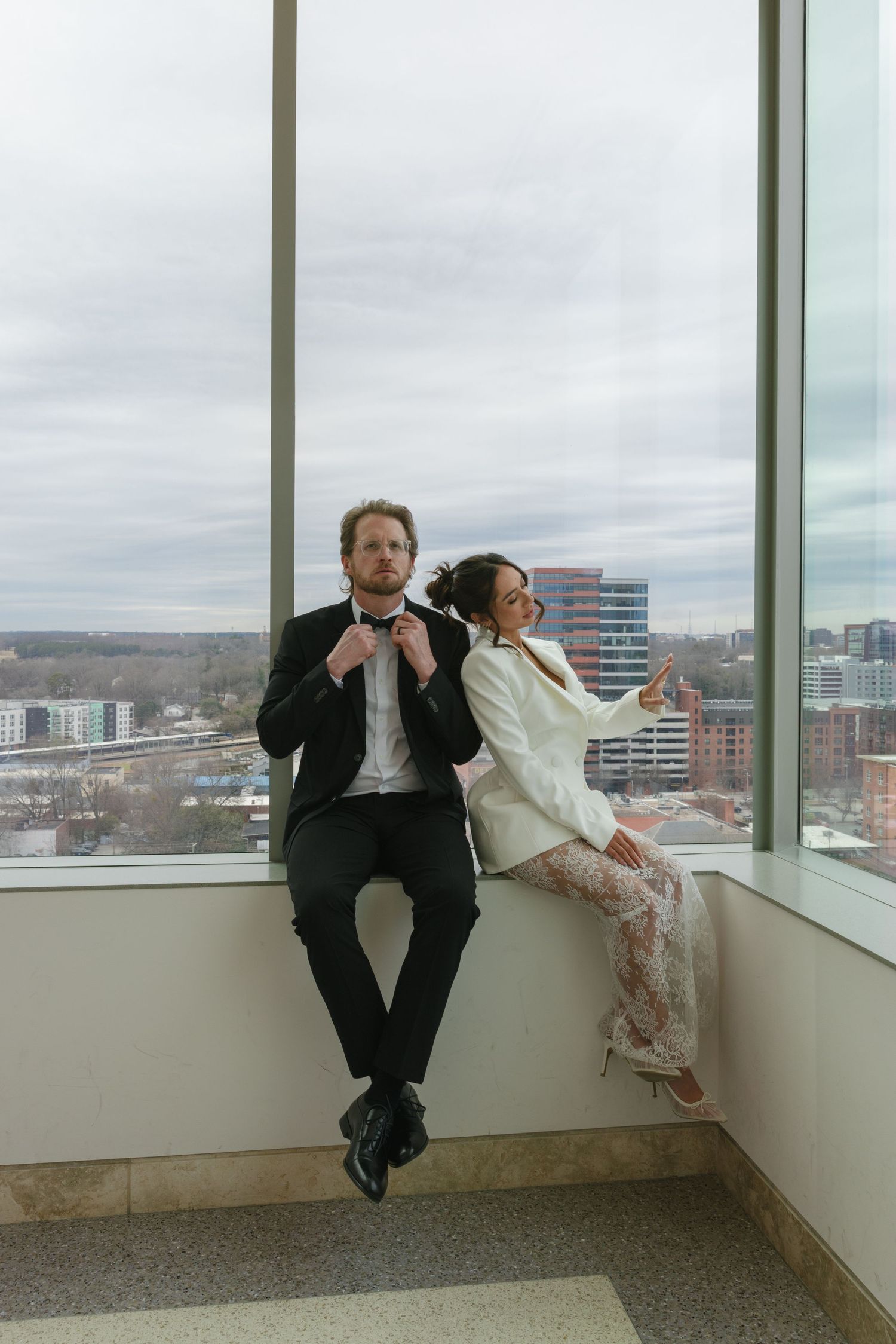 A couple in formal attire sits on a window ledge with a city skyline backdrop, man adjusting bow tie, woman in white blazer.