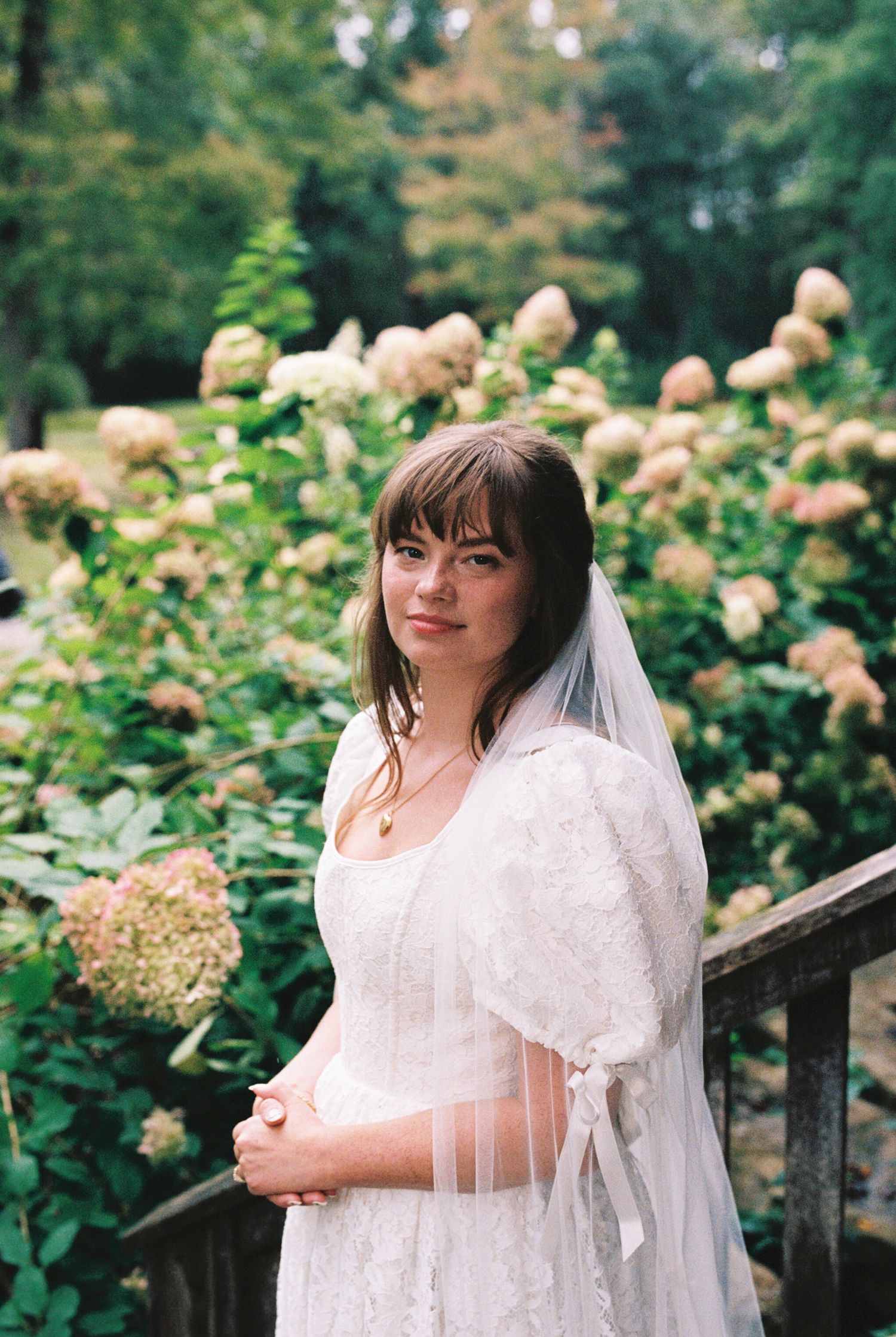 A bride in a white lace dress and veil poses in a garden filled with blooming hydrangea flowers on a summer day.