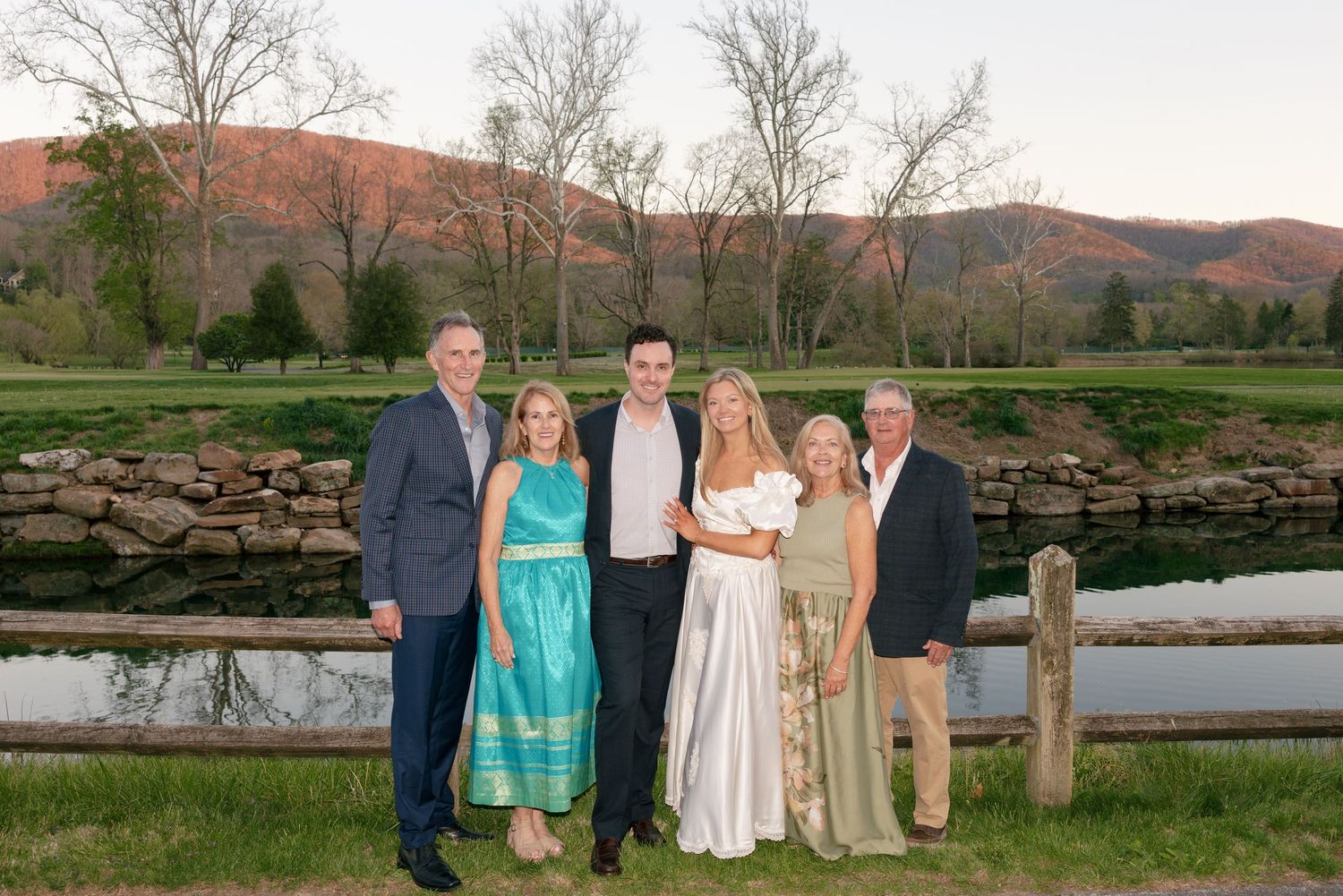 Family group photo outdoors at sunset with rolling green hills and a pond in the background.