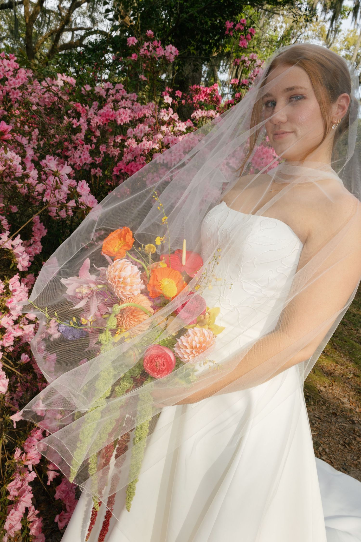 Bride in white strapless gown and veil holds colorful bouquet outdoors near pink flowering bushes.