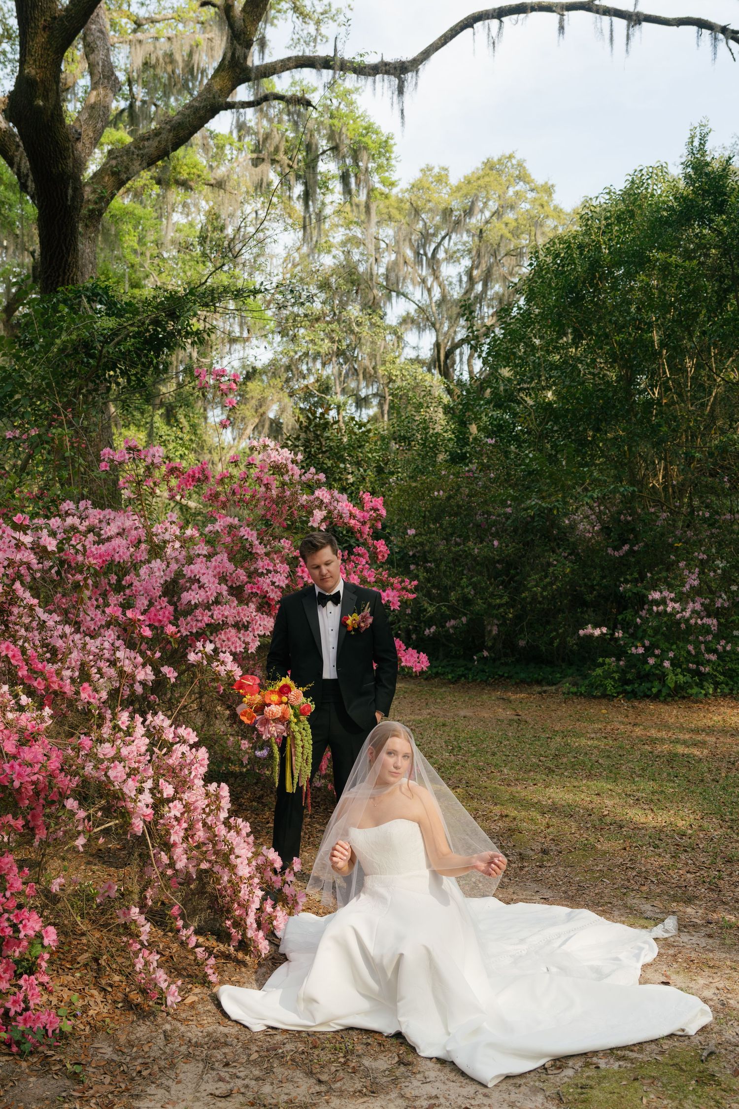 Bride in white gown sits on ground near pink azaleas as groom in tuxedo stands behind in lush garden.
