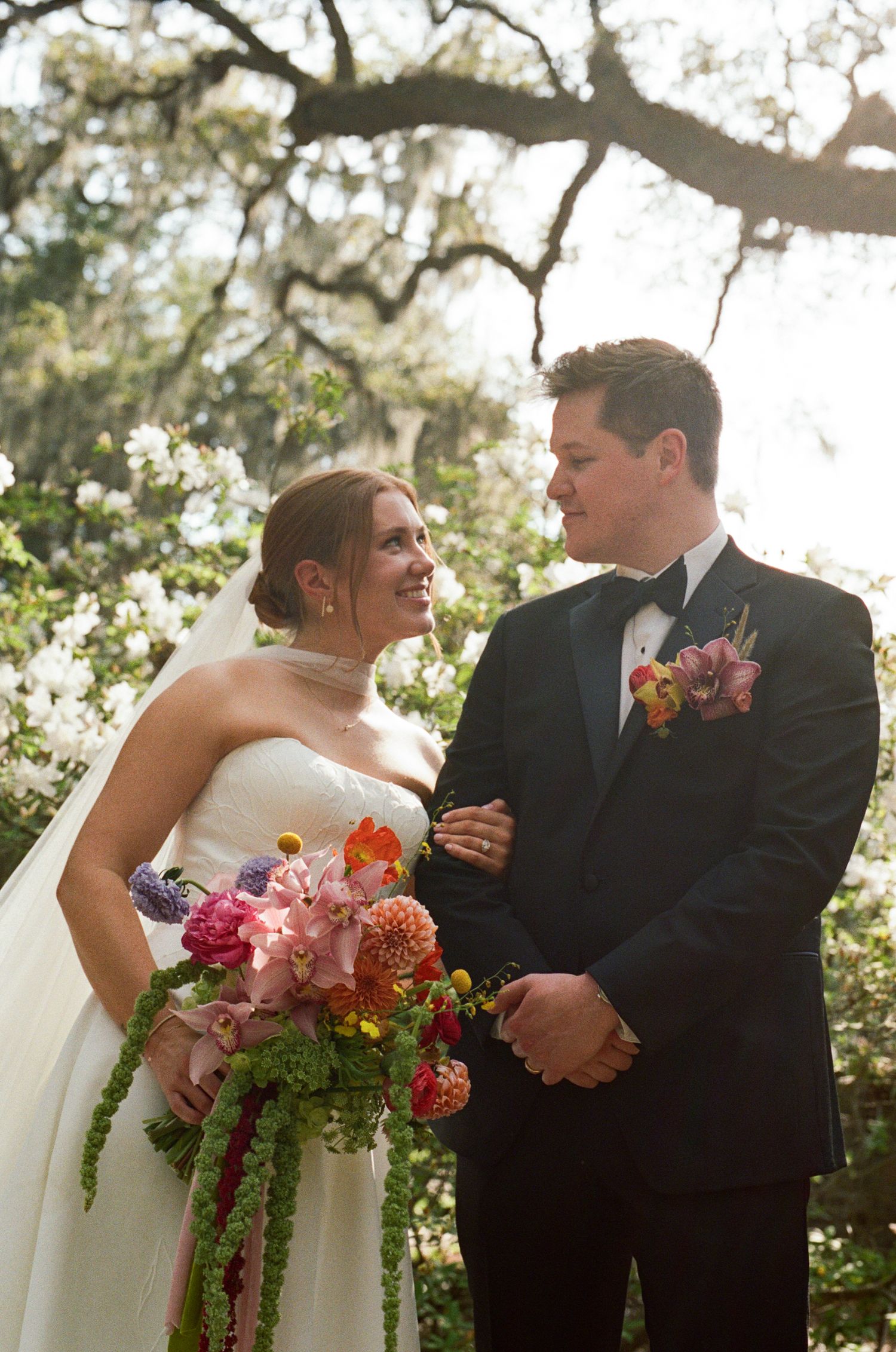 Bride and groom share a joyful moment outdoors, she holds a vibrant bouquet of pink and orange flowers under moss-draped trees.