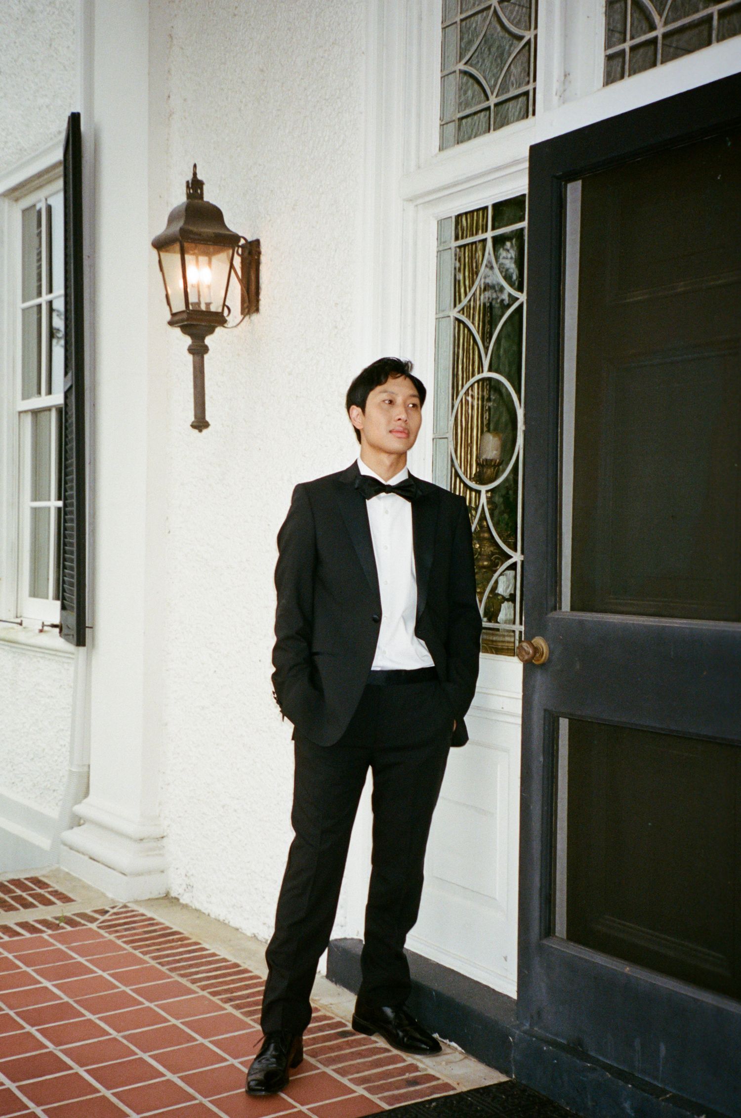 A person in a black tuxedo and bow tie poses in front of a white colonial-style building entrance.