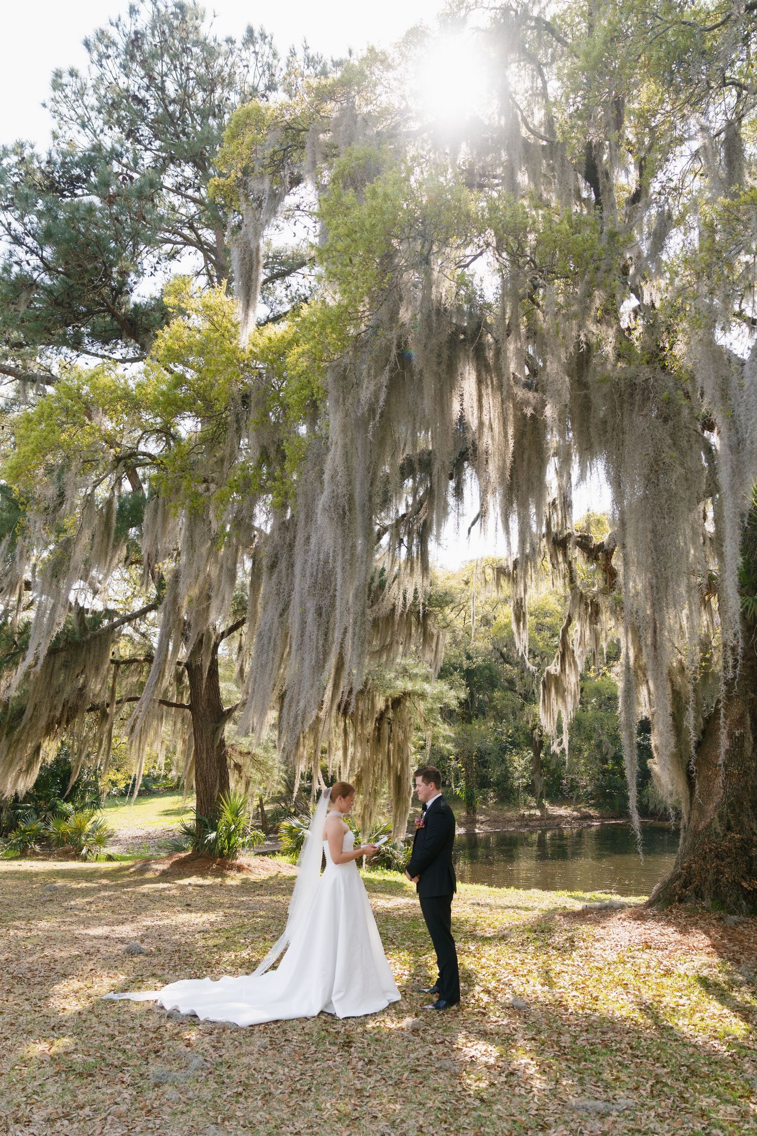 Bride and groom share a romantic first look under Spanish moss-draped oak trees beside a serene pond in a Southern garden.