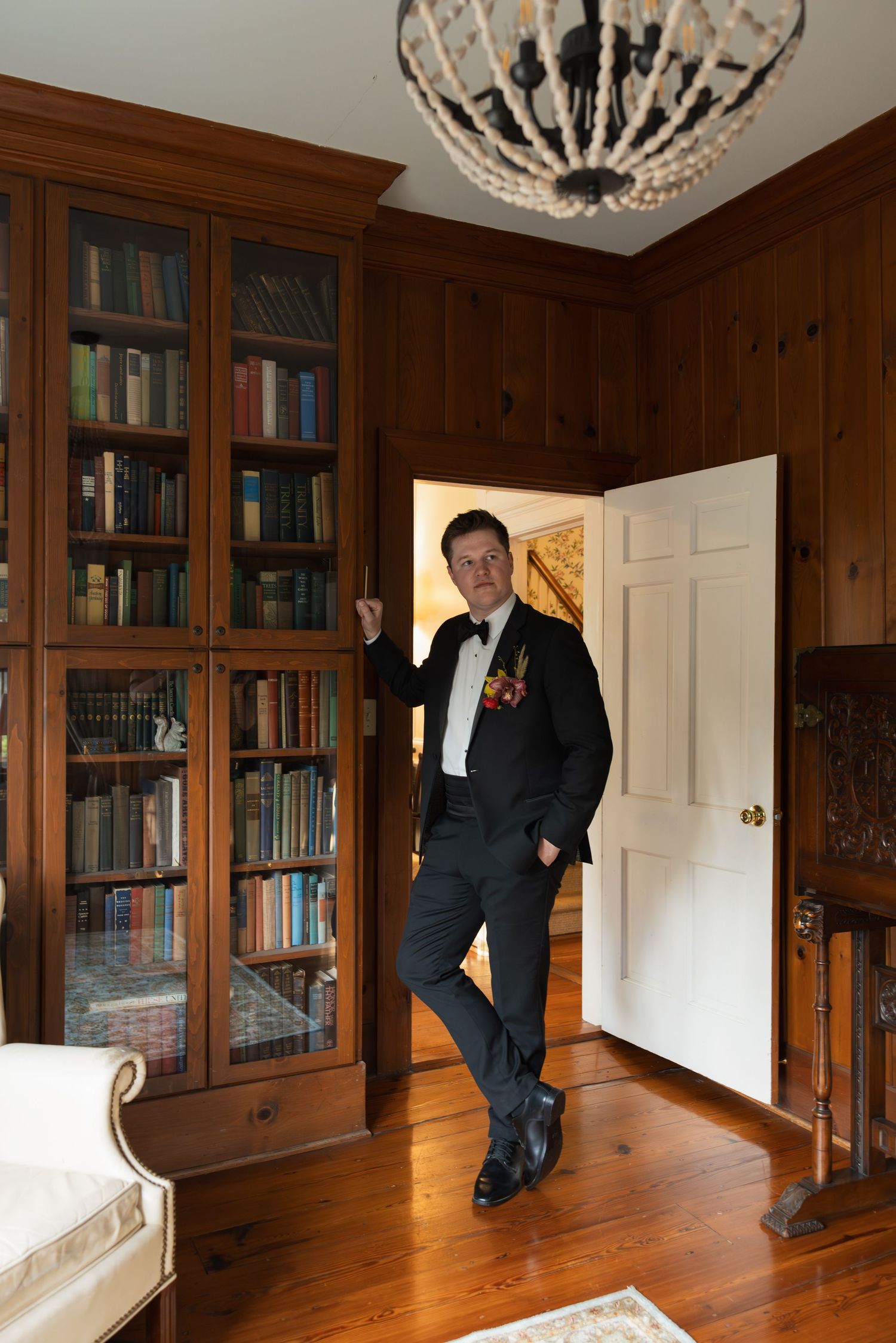 Groom in tuxedo with boutonniere poses in doorway of elegant wood-paneled library with crystal chandelier.