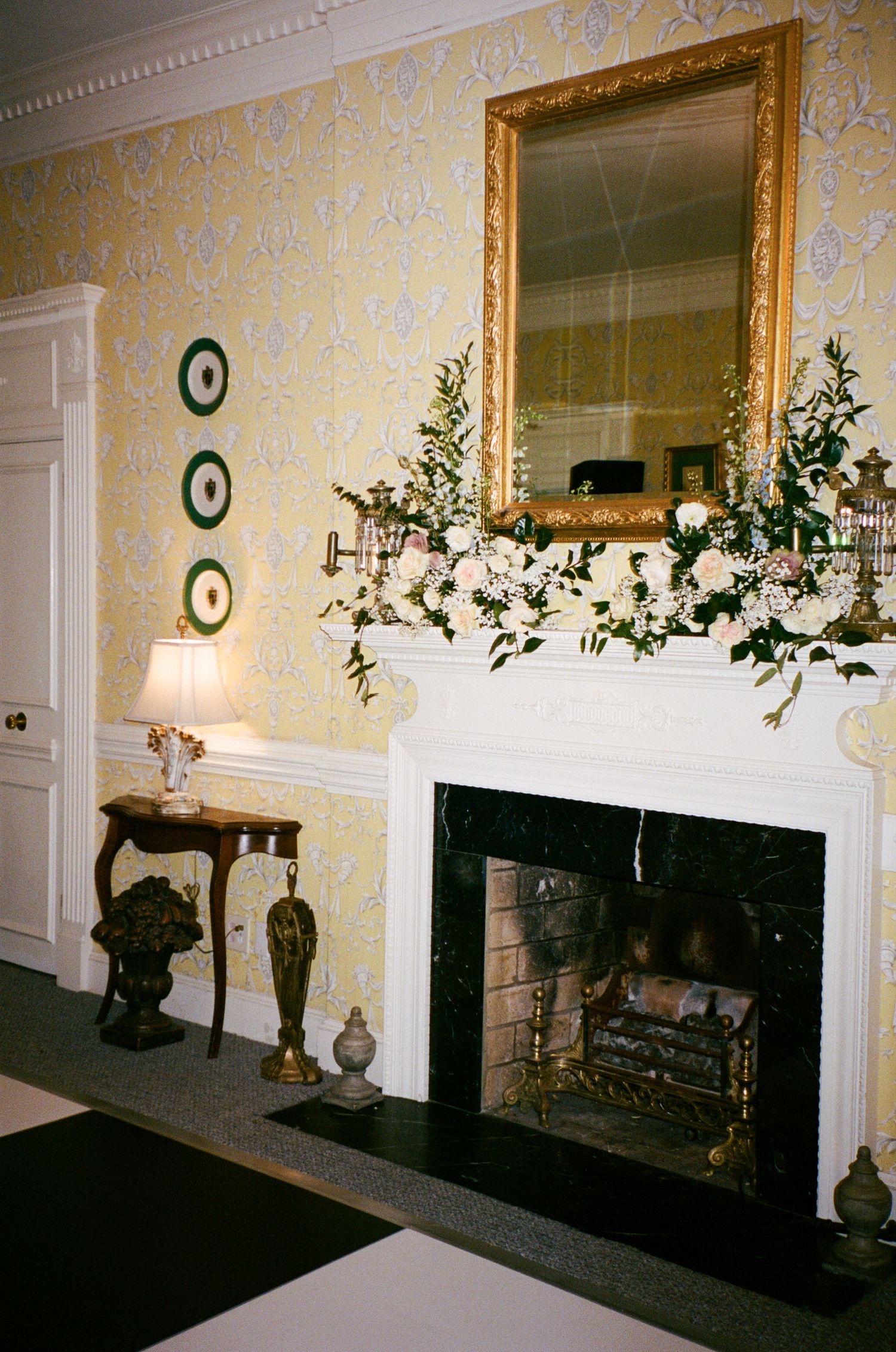 Elegant fireplace with white mantel decorated with pink and white floral garland, gold framed mirror, and yellow damask wallpaper.