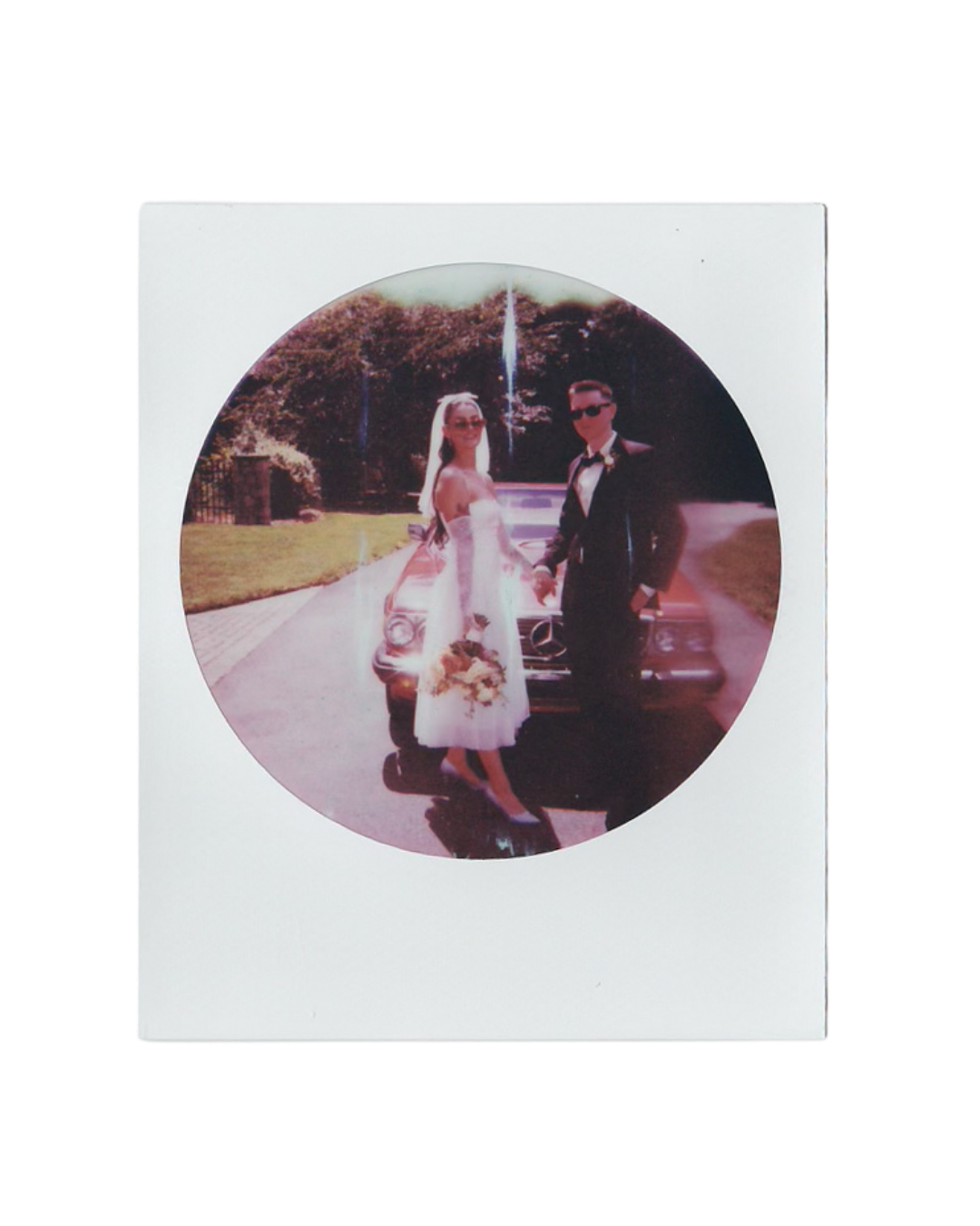 Bride in a white dress and veil holding a bouquet stands beside a groom in a tuxedo and sunglasses in front of a vintage car in a round inst