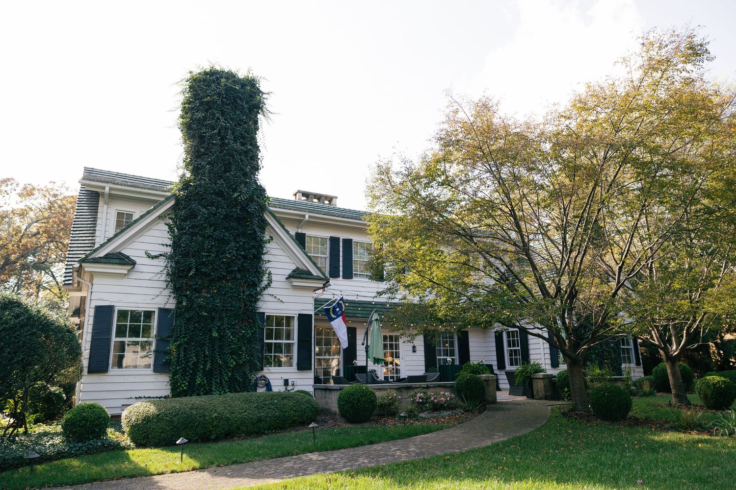 White two-story colonial house with black shutters, an American flag, ivy-covered chimney, manicured shrubs and a curved brick walkway.