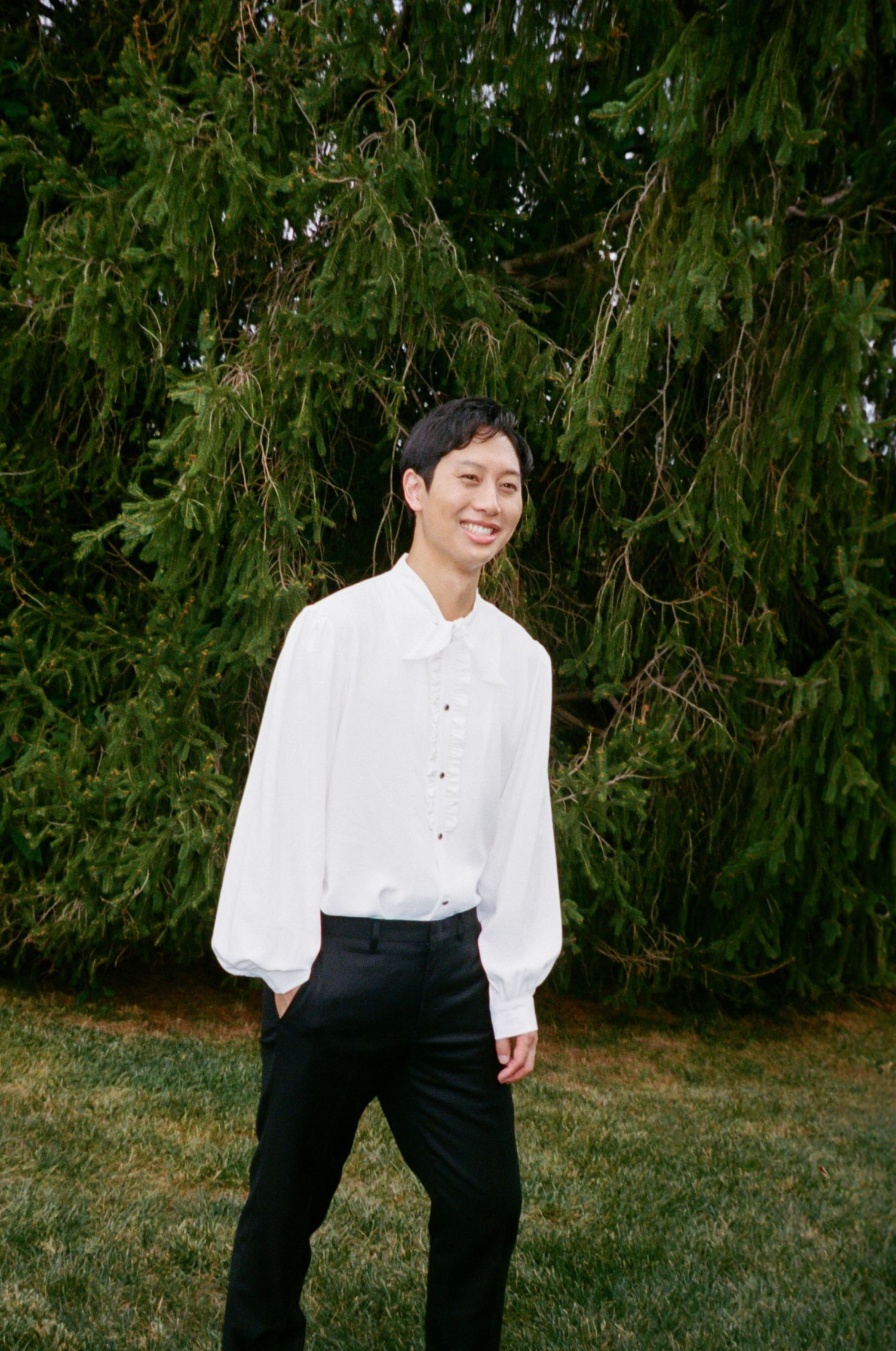 A person in a white button-down shirt and black pants stands smiling in front of an evergreen tree.