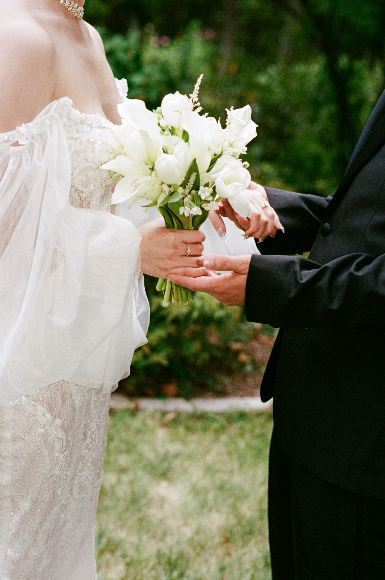 Close-up view of hands exchanging wedding rings during an outdoor ceremony with white floral bouquet and lace dress details.