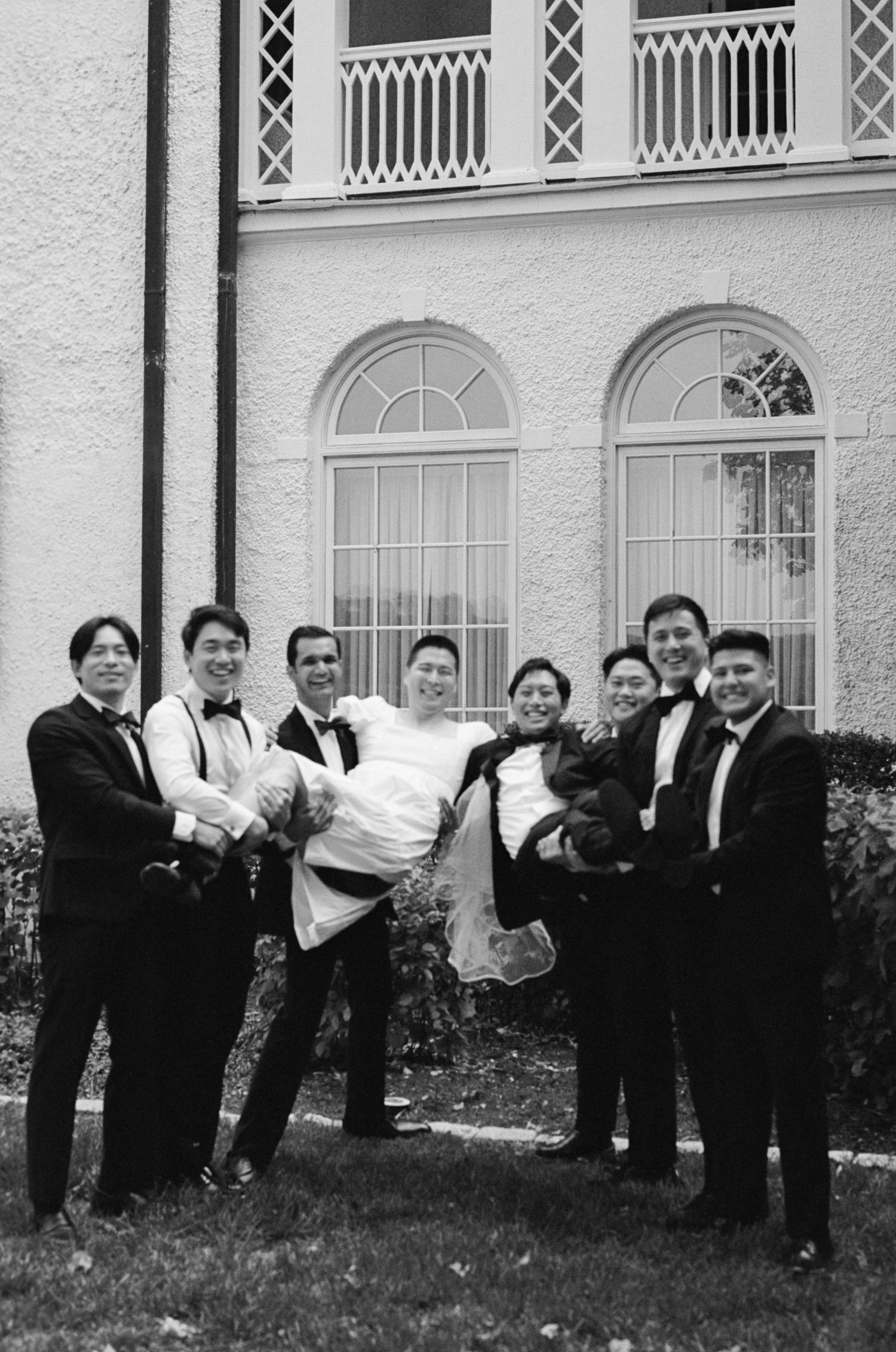 A fun wedding party group poses laying sideways on grass outside an elegant white building with arched windows.