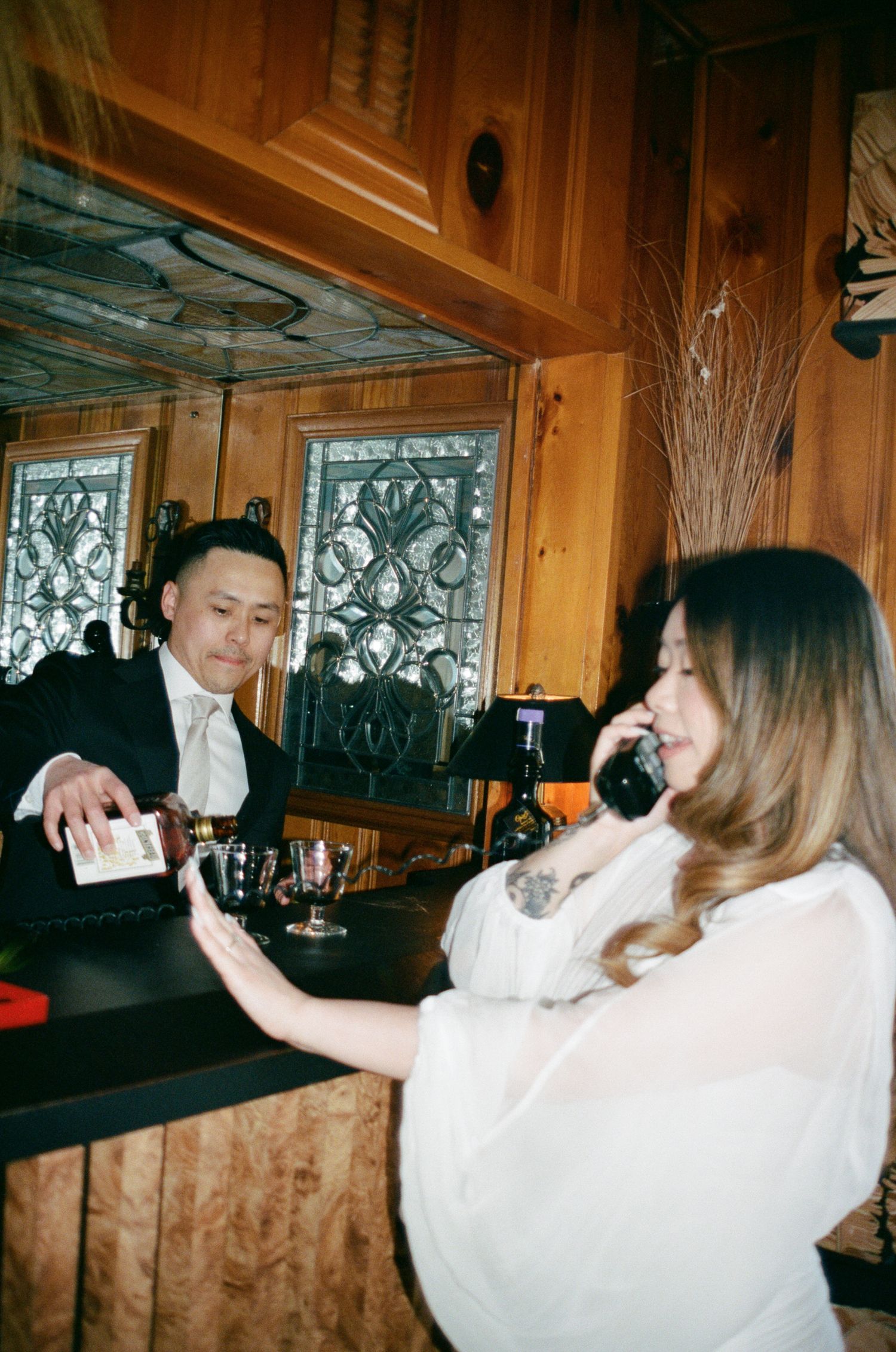 A bartender pours wine for a customer at a rustic bar with ornate glass windows in the background.