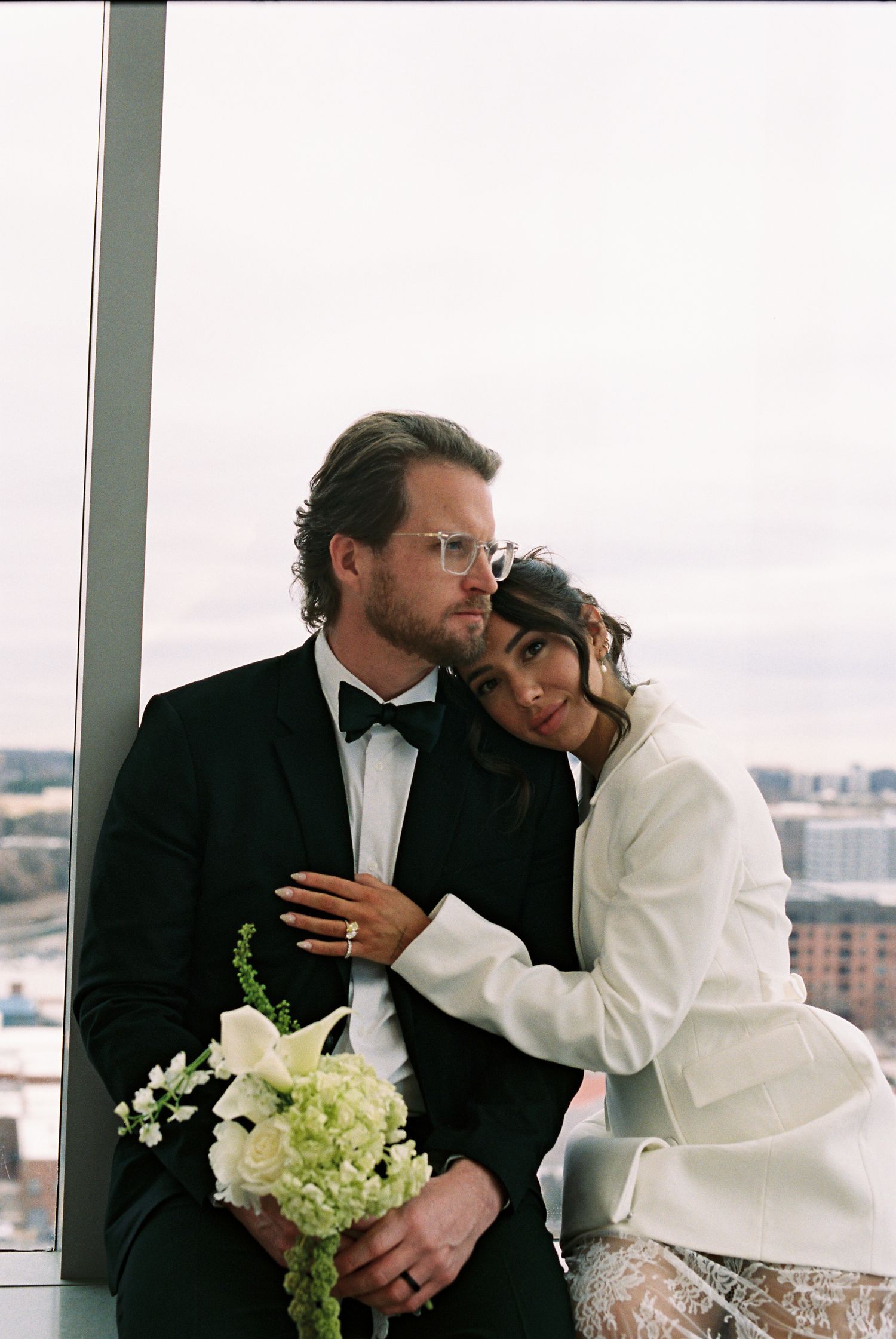 Elegant bride and groom in black tuxedo and white blazer pose by floor-to-ceiling windows holding white floral bouquet.