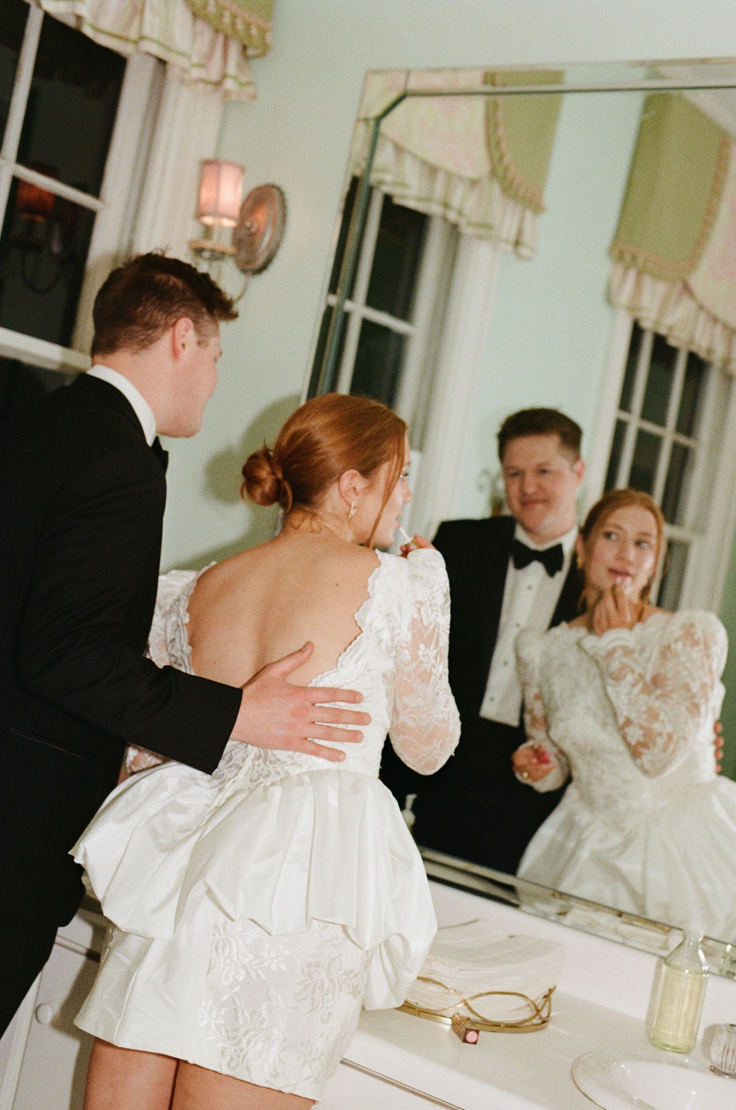 Bride in short lace wedding dress with bow bustle stands with groom in tuxedo, reflected in vintage mirror.