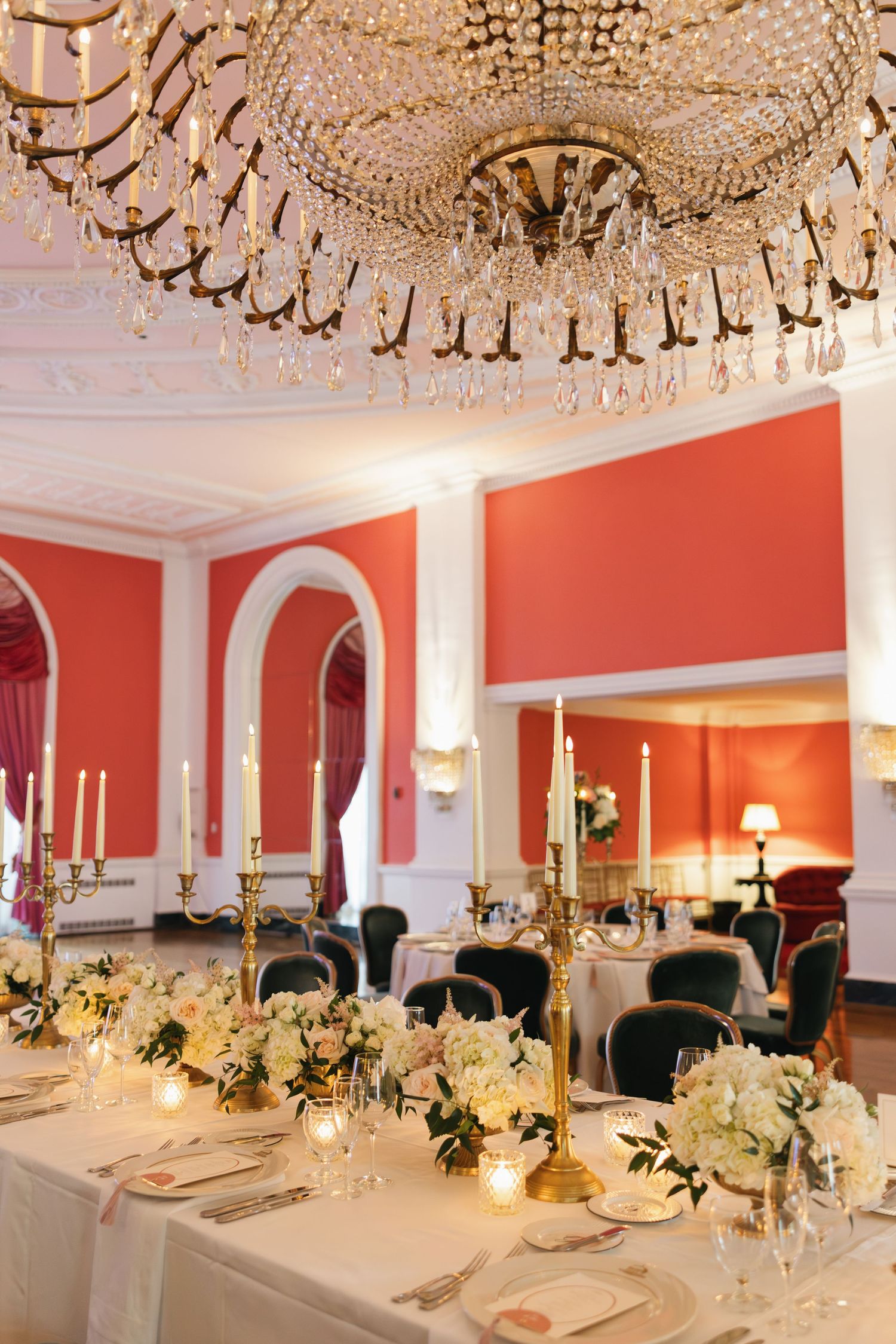 Elegant wedding reception in a red ballroom with crystal chandelier, gold candelabras, and white floral centerpieces.