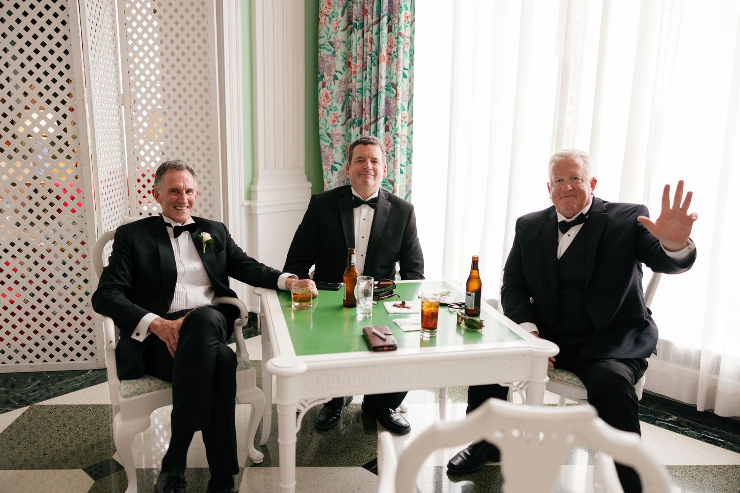Three men in tuxedos sitting at a white table with beer bottles, smiling and waving at a formal event.