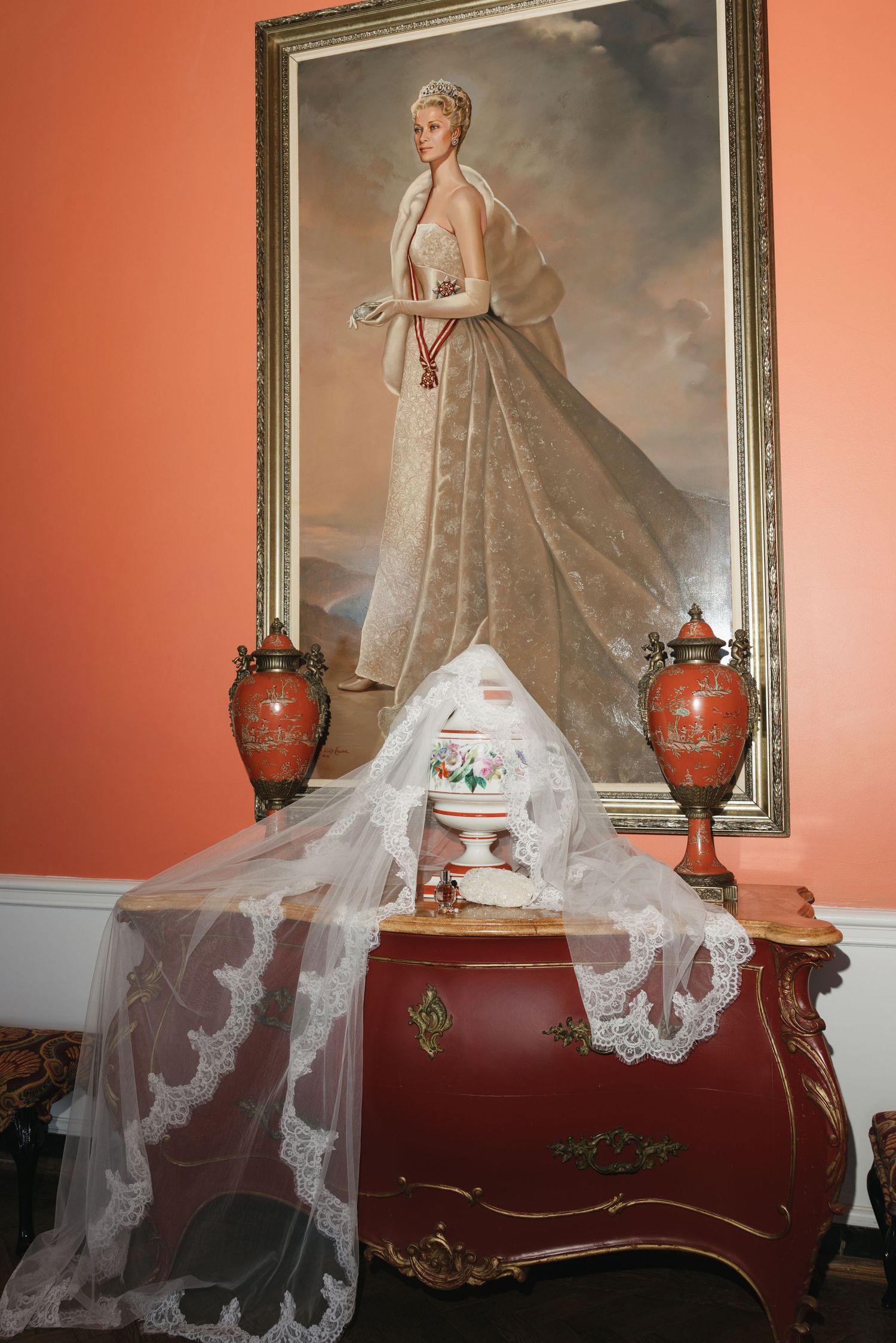 Elegant bridal veil draped over ornate red dresser beneath portrait painting in coral-walled room with decorative urns.