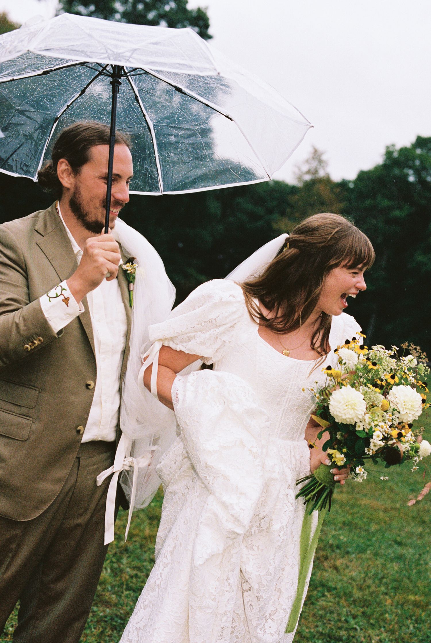 A couple shares a joyful moment under clear umbrellas during their rainy day wedding celebration in a green field.