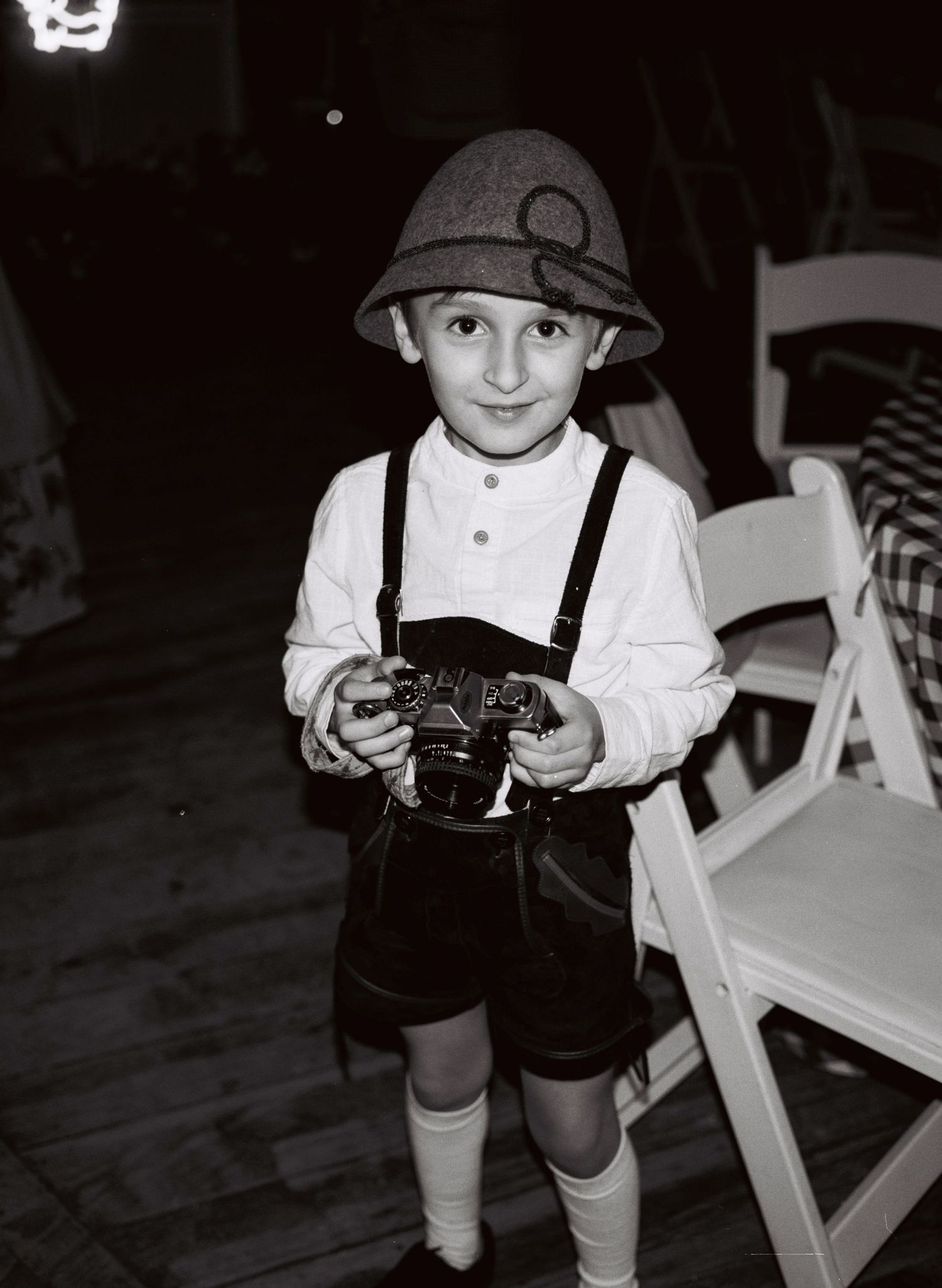 Vintage black and white photo of a child in lederhosen, suspenders and a helmet holding an old camera.