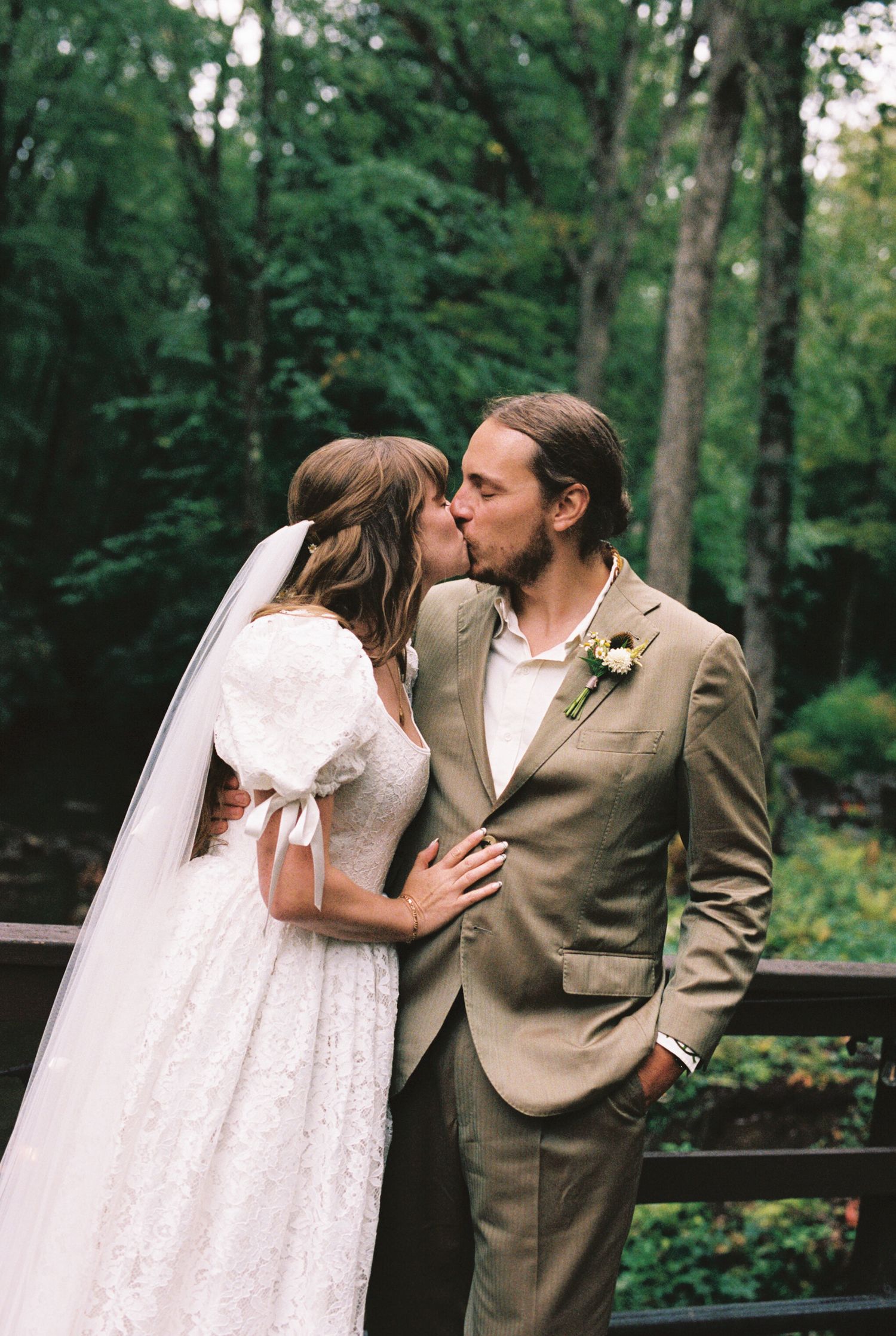 A romantic wedding moment captured in a lush forest setting with a bride in white lace and groom in tan suit.