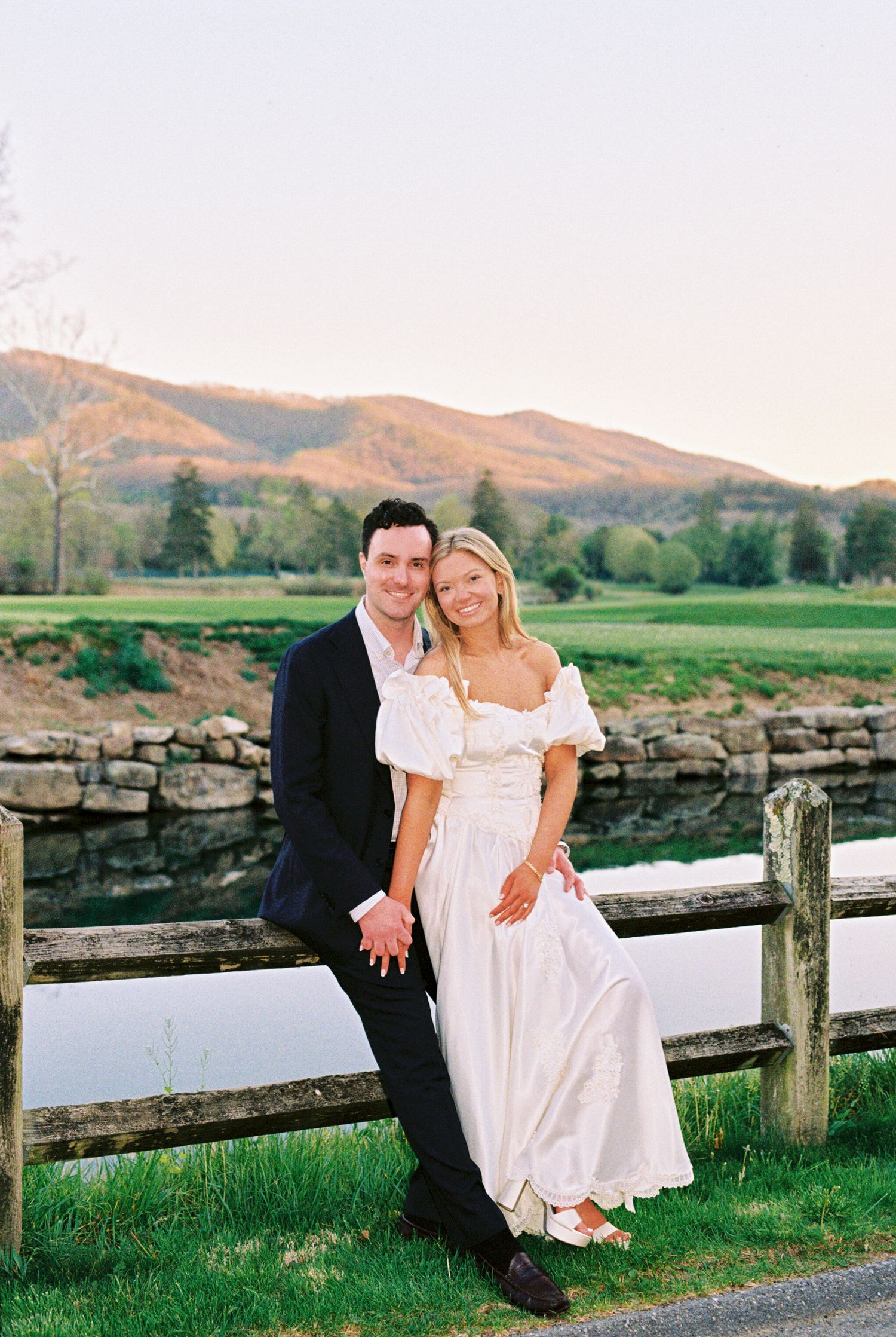 Couple posing by a wooden fence with a scenic mountain backdrop and pond during golden hour.