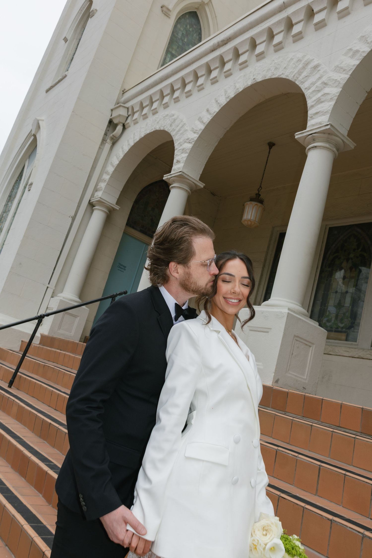 Bride in white blazer dress and groom in black tuxedo kissing on church steps on their wedding day.