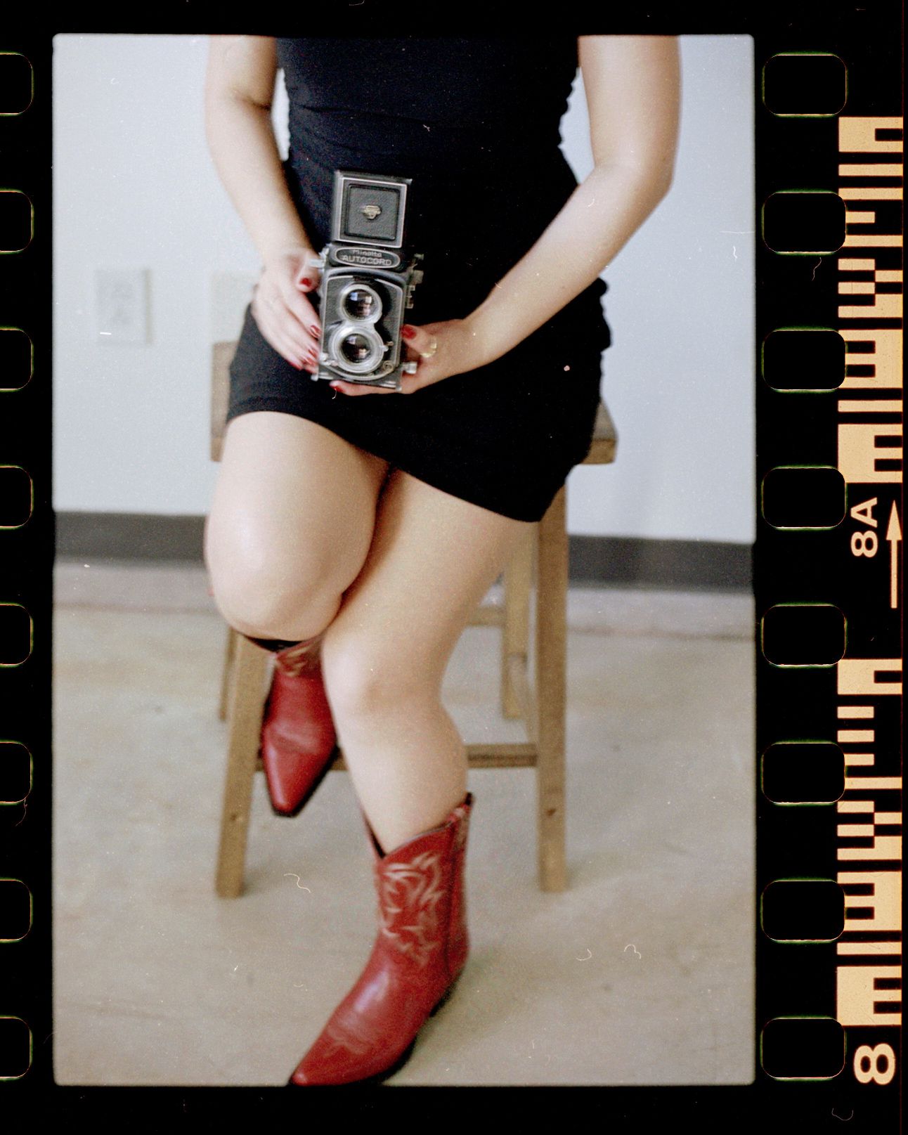 Woman in black dress and red cowboy boots holds a vintage twin-lens reflex camera while sitting on a wooden stool.