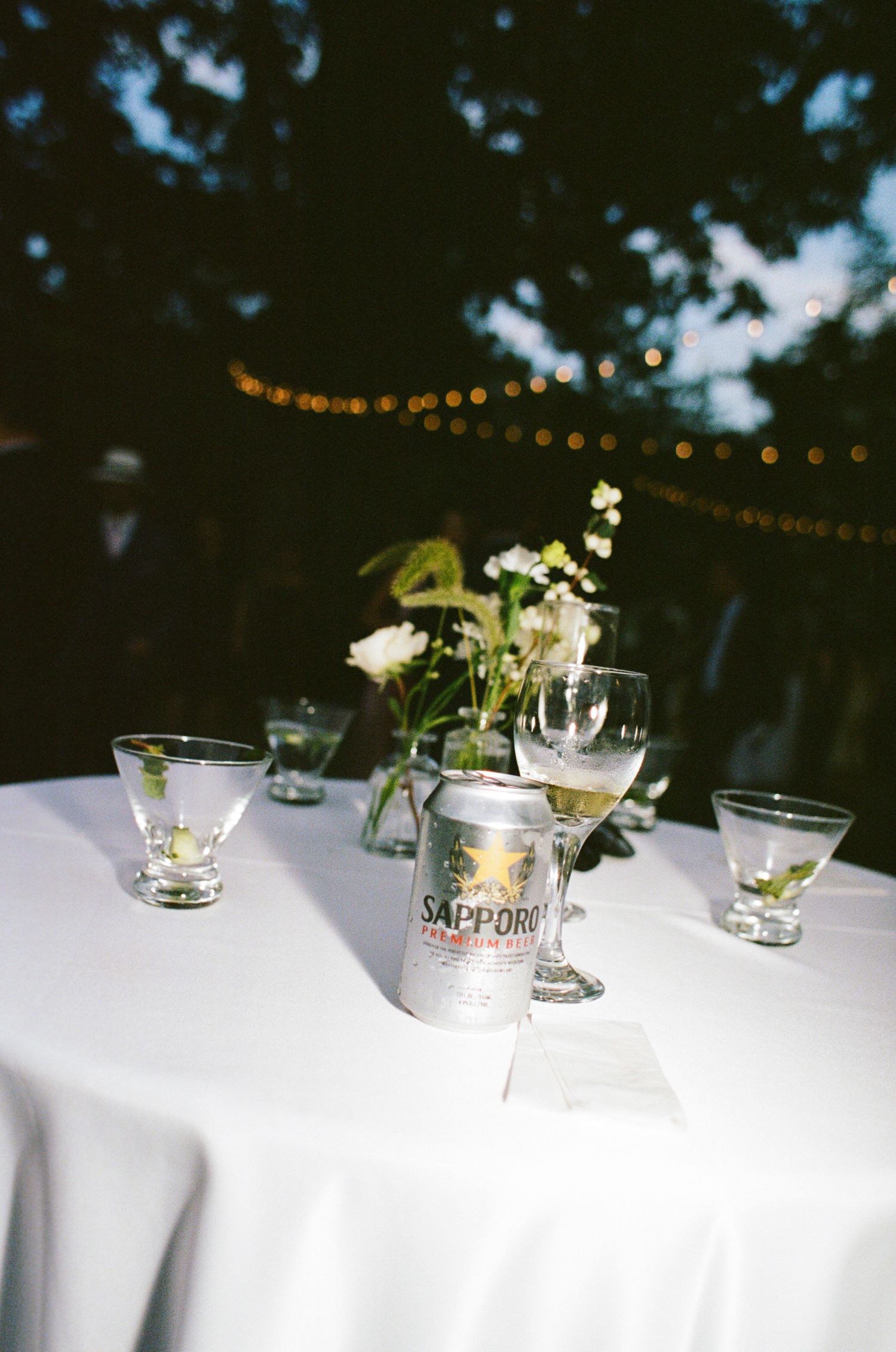 String lights illuminate white tablecloths with delicate floral centerpieces and glassware at an elegant outdoor event.
