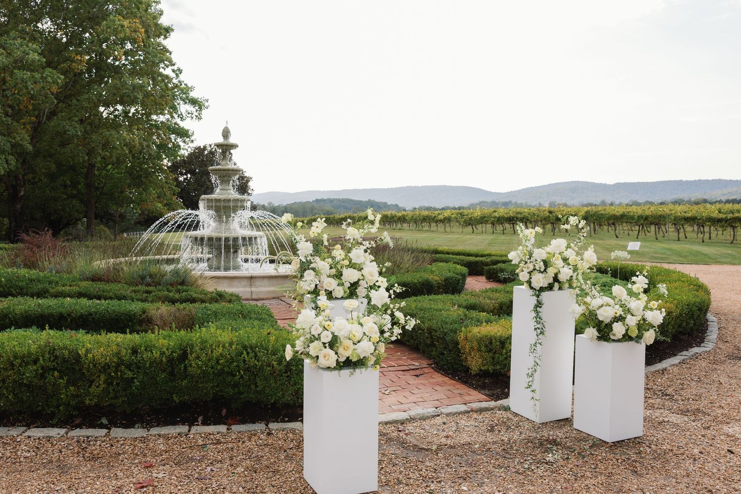 White floral arrangements in elegant vases line a rustic outdoor pathway with vineyard and mountain views in the background.