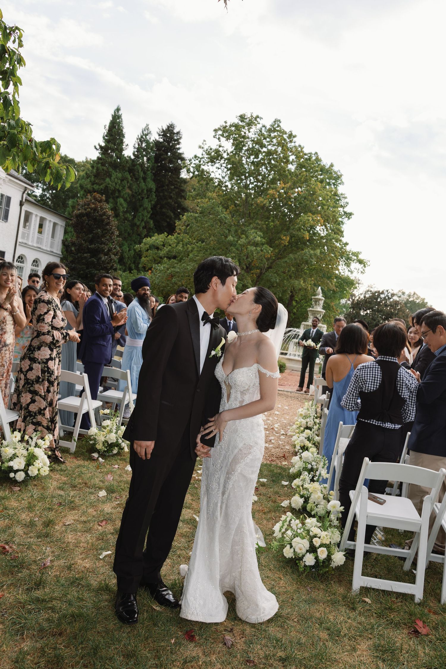 A romantic outdoor wedding ceremony with guests seated on white chairs and floral decorations along a garden path.
