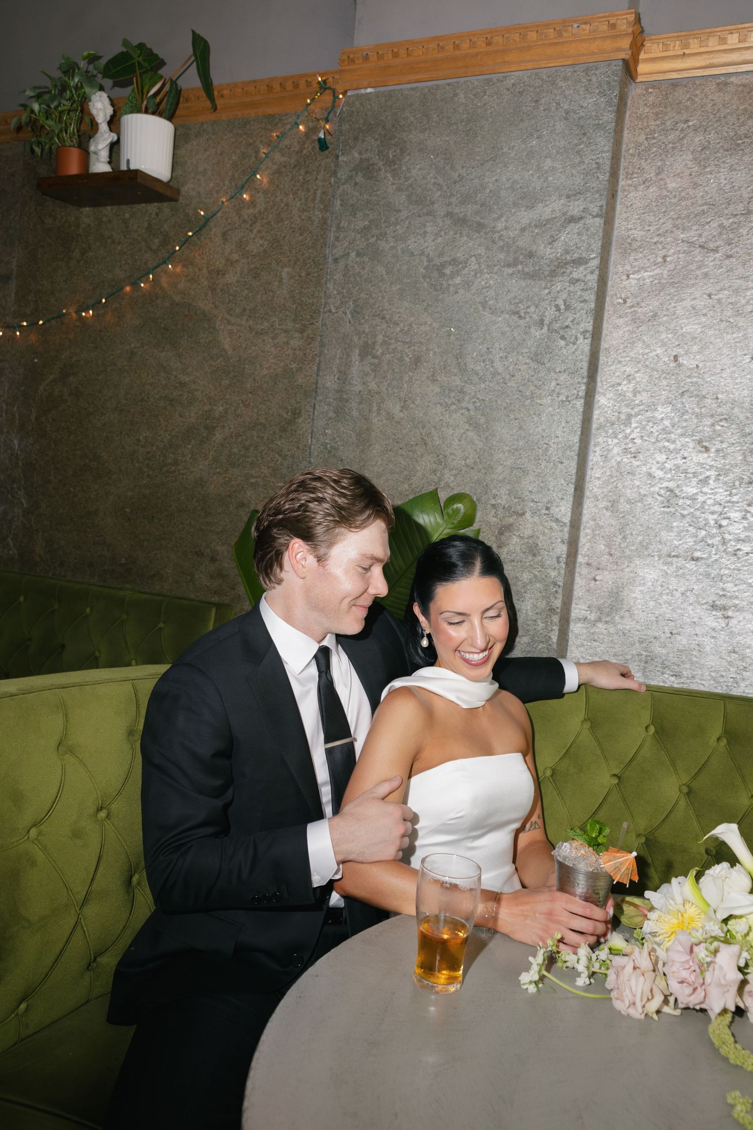 Bride and groom sitting together on a green velvet booth, smiling and holding drinks at their wedding reception.