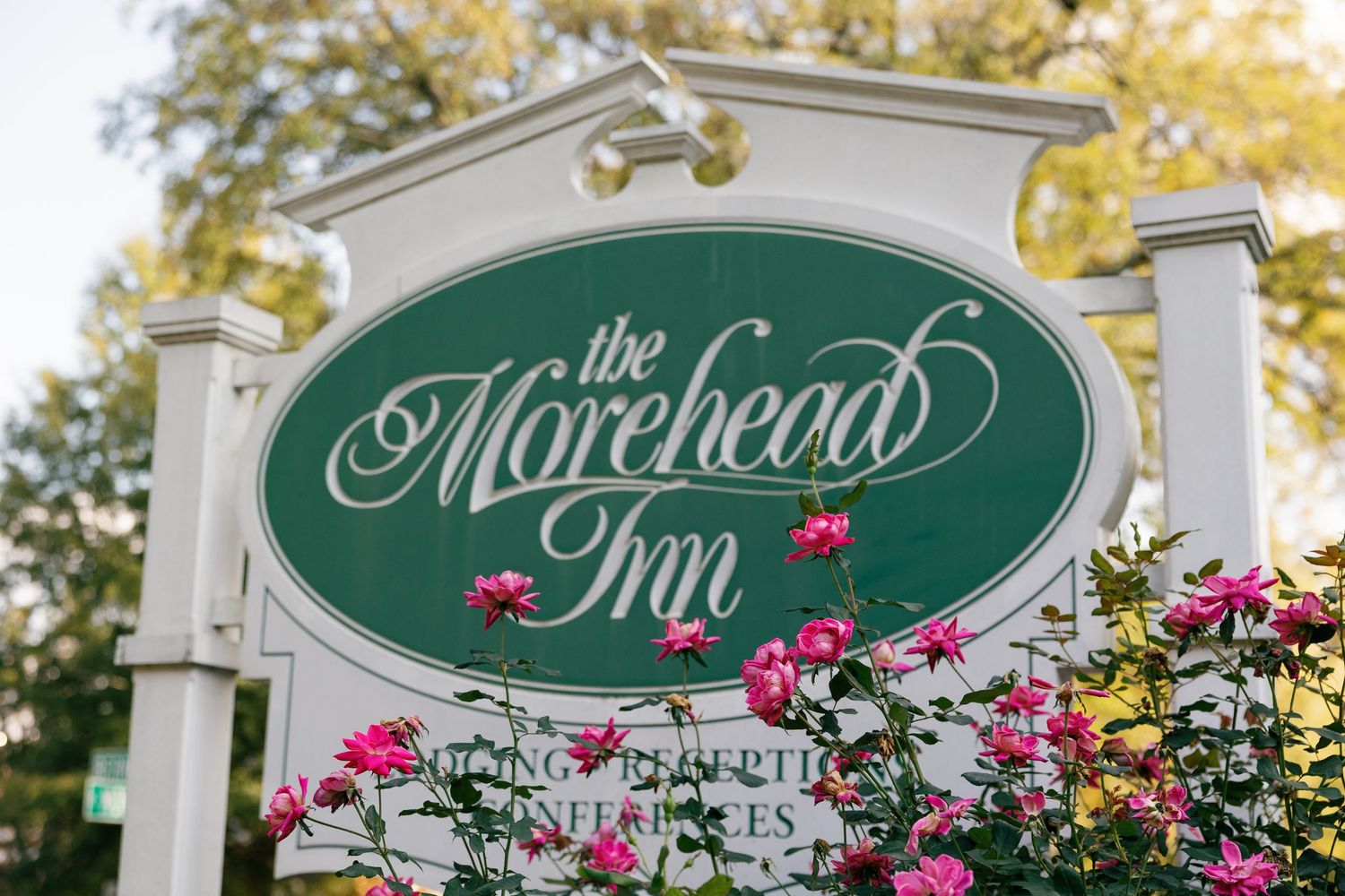 Green and white sign reading The Morehead Inn with pink roses blooming in the foreground and autumn trees behind.