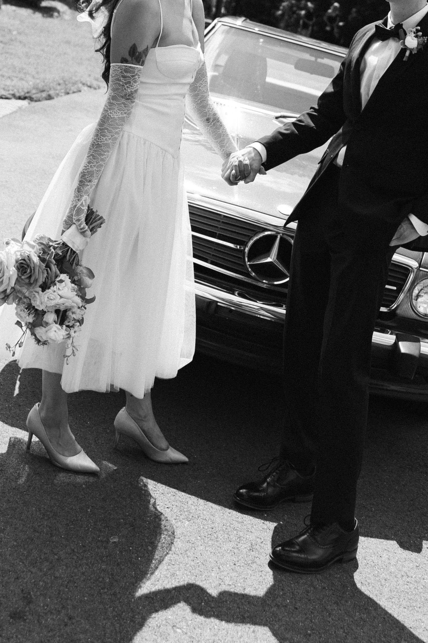 Black and white photo of a bride in a lace-gloved tulle dress holding a bouquet and the groom's hand beside a vintage Mercedes-Benz.