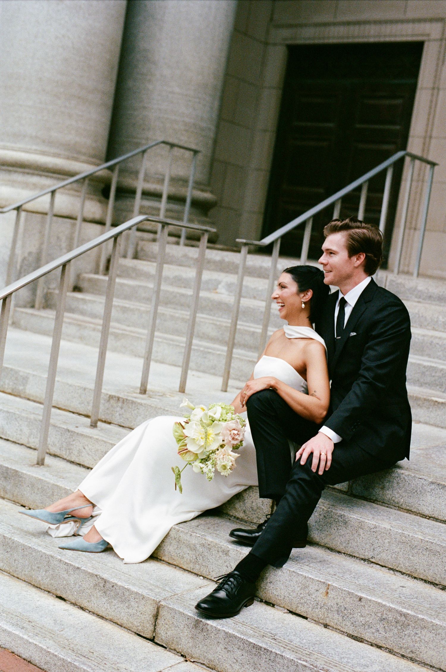 Bride in a white strapless gown holding a bouquet sits beside a groom in a black suit on stone steps outside a classic columned building.