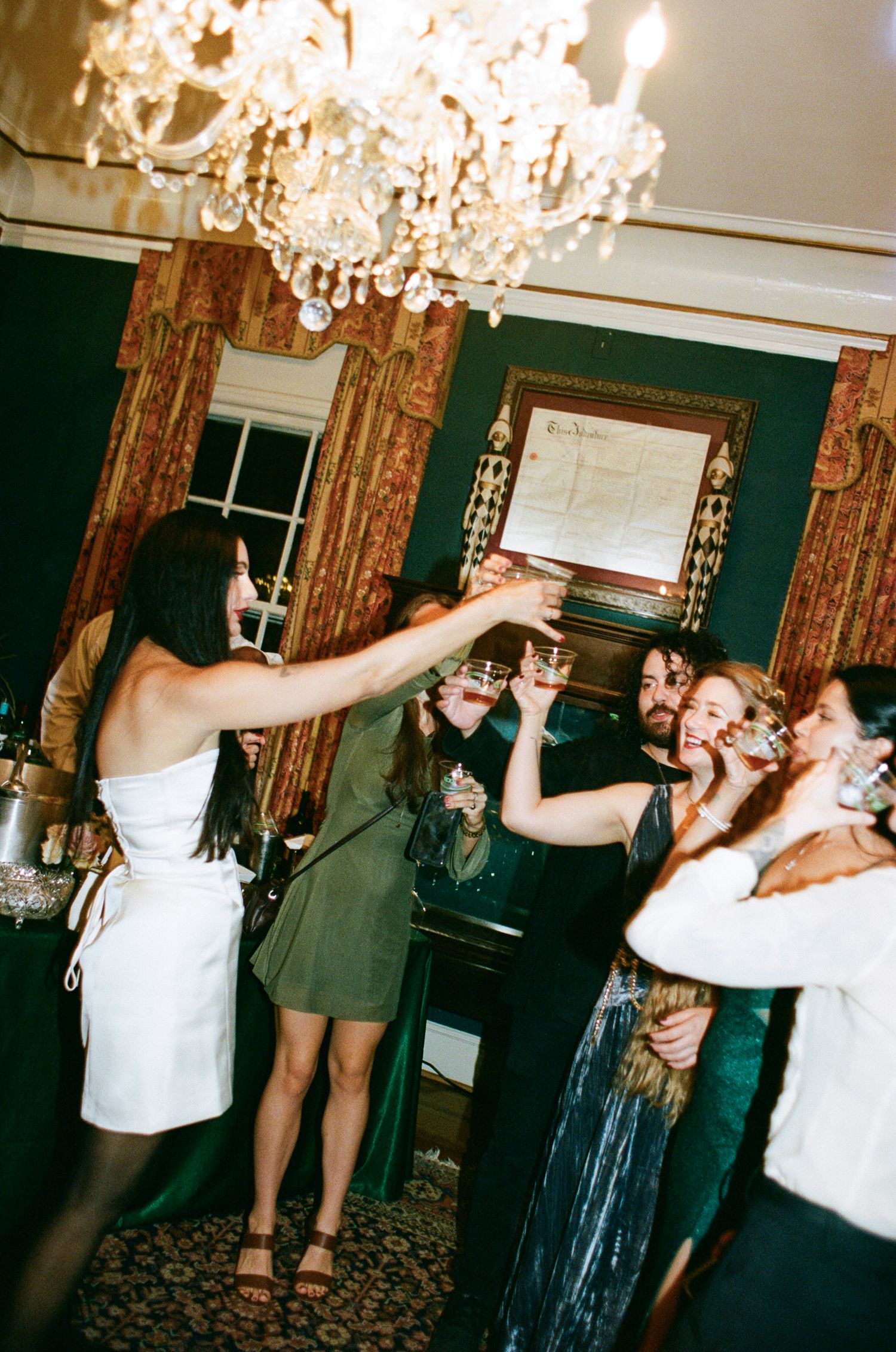 Guests in elegant attire raise glasses in a festive toast beneath a crystal chandelier in an ornate room with green walls and gold drapes.