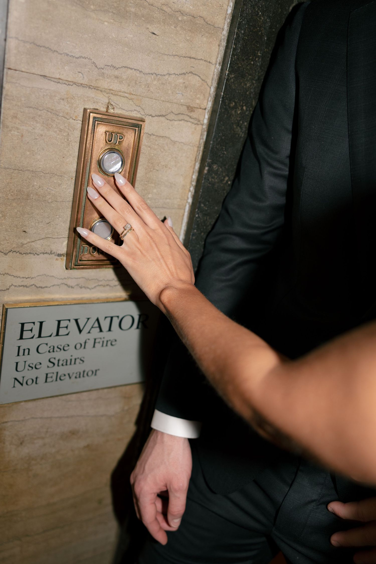 A woman's manicured hand with a ring presses the up button beside an elevator fire safety sign as a man in a dark suit stands nearby.