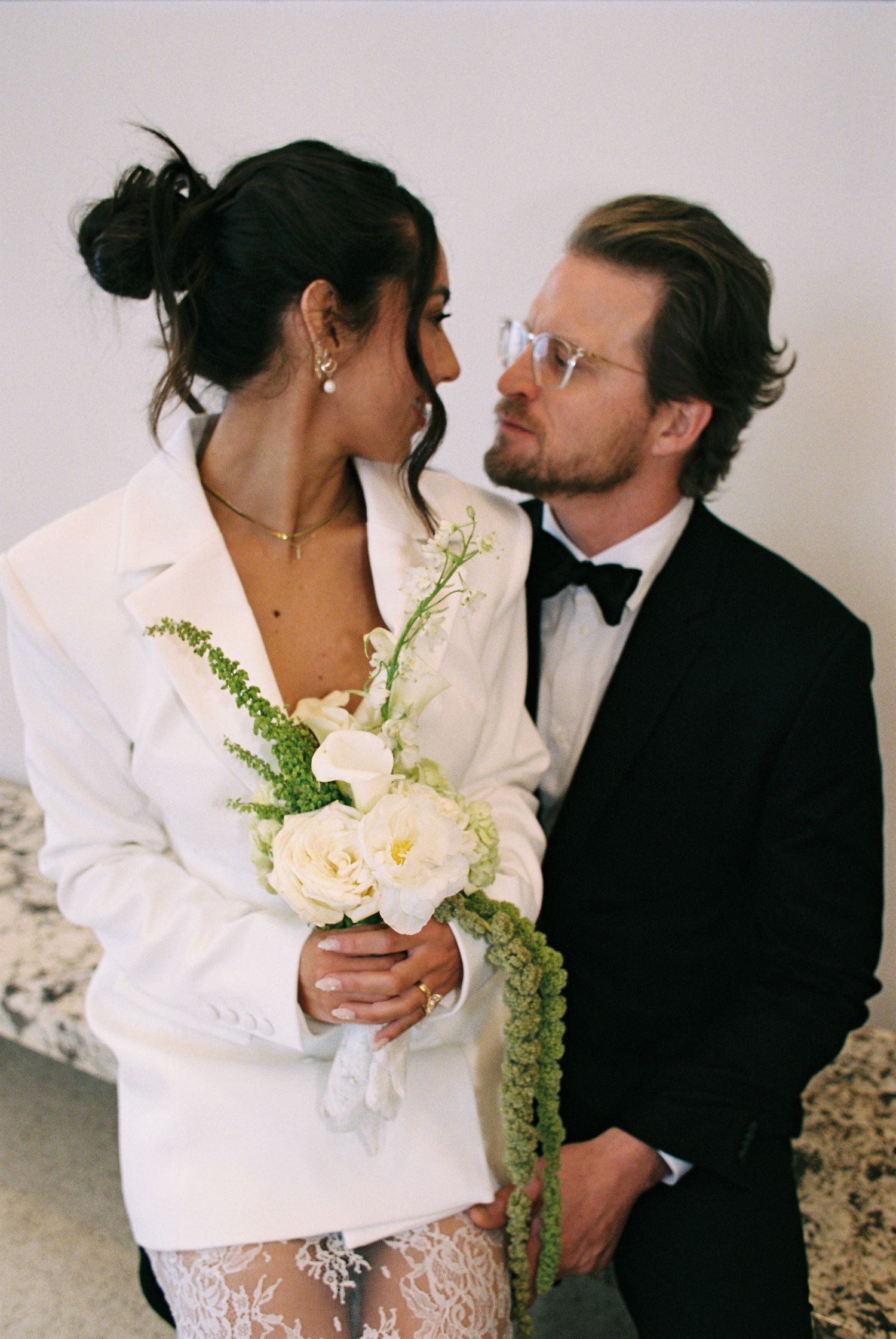 Couple in wedding attire, woman in white blazer with bouquet, man in black tuxedo, gazing at each other.