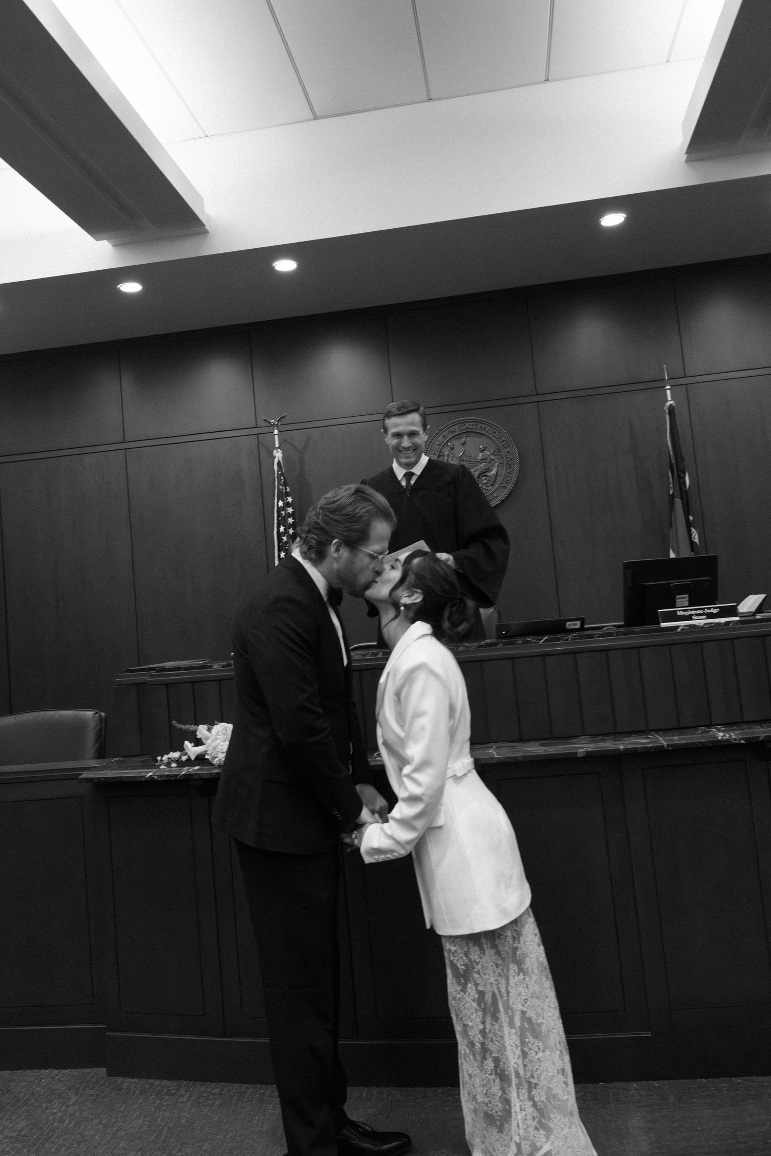 Black and white photo of a couple kissing after a courthouse wedding ceremony, with a smiling judge presiding in the background.