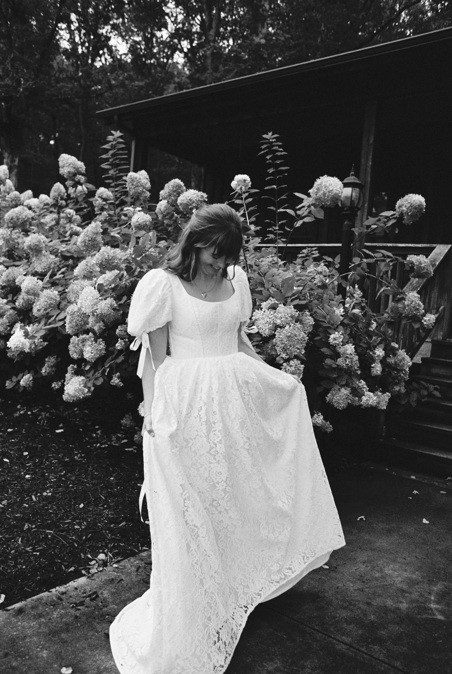 A flowing white lace dress hangs suspended in a garden with blooming hydrangea bushes in the background.