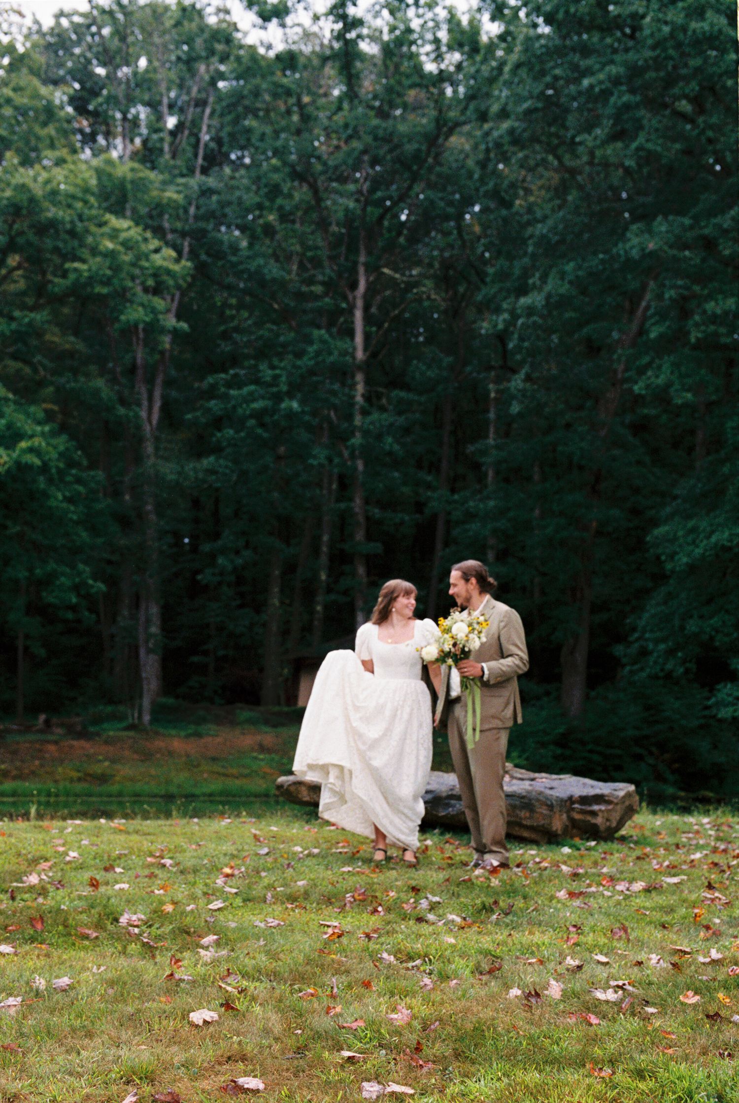 A couple sits together on a grassy hillside surrounded by lush green trees in a romantic outdoor setting.